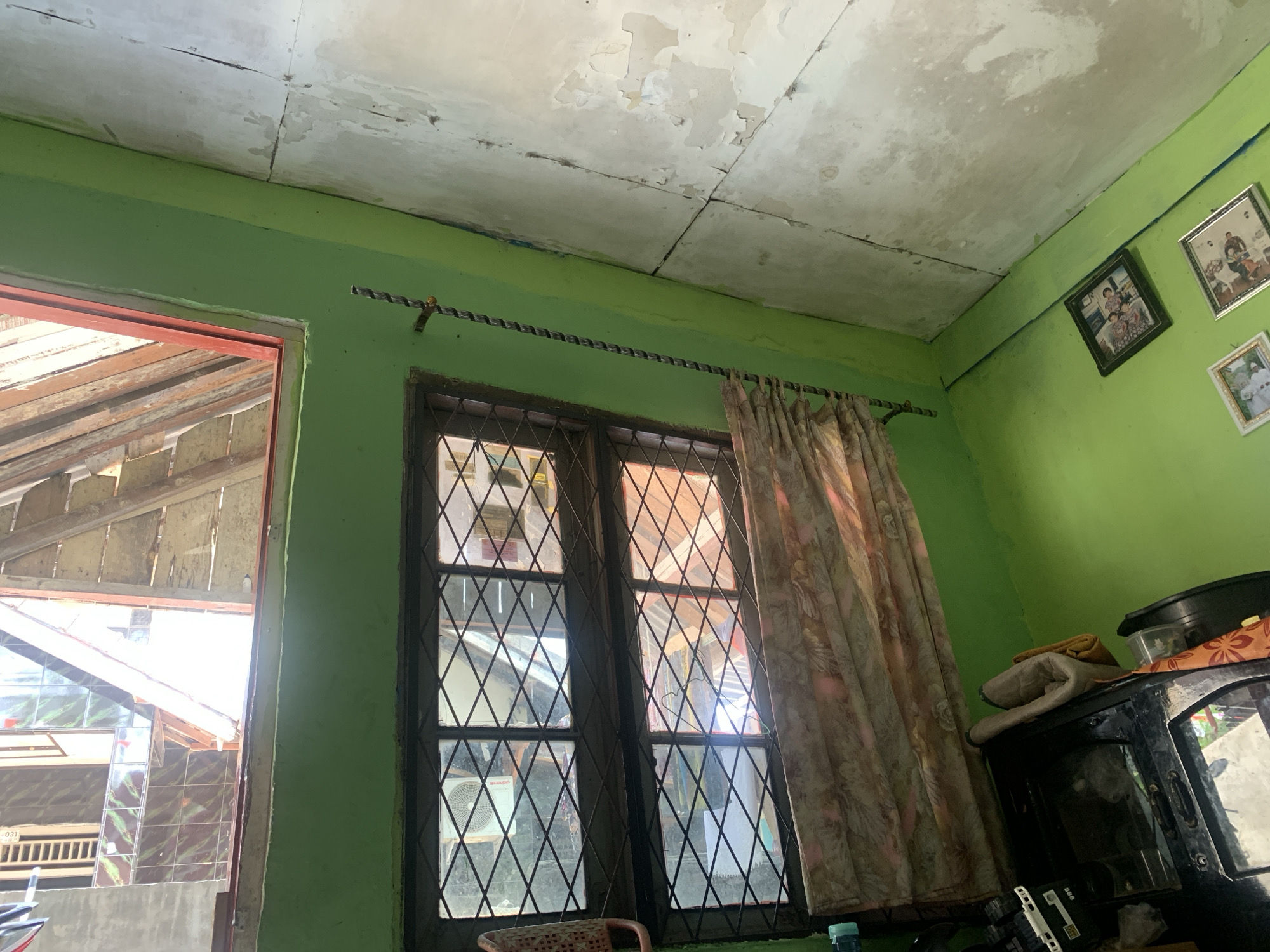 This image depicts an interior room with bright green walls and a damaged ceiling in Palembang, Indonesia. The ceiling shows signs of peeling paint and water damage, with visible cracks and stains. A window with a diamond-patterned metal grate is present, covered partially by a floral-patterned curtain. Outside the window, wooden structures and glimpses of other buildings are visible. On the green wall, there are several framed photographs displayed, showing people. To the right of the window, a black cabinet or furniture piece is visible, with some folded fabric and containers on top. The overall impression is of a modest, lived-in space in a warm climate.