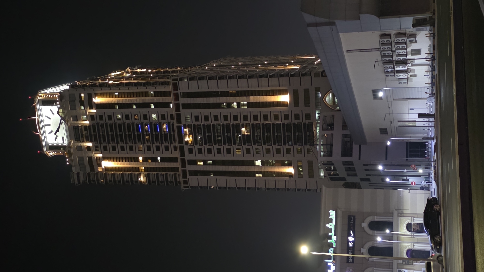 The image shows a tall, modern skyscraper at night in Ajman, United Arab Emirates. The skyscraper has a large clock face at its summit and is illuminated with warm yellow lights along its vertical accents. Below the skyscraper, there is a lower-rise building with a sign that reads "سفير هايب" in Arabic and "SHOES US". A dark-colored sedan is parked in front of this building, and streetlights illuminate the scene. The sky is dark, indicating it is nighttime. The building's architecture suggests a commercial or residential purpose.