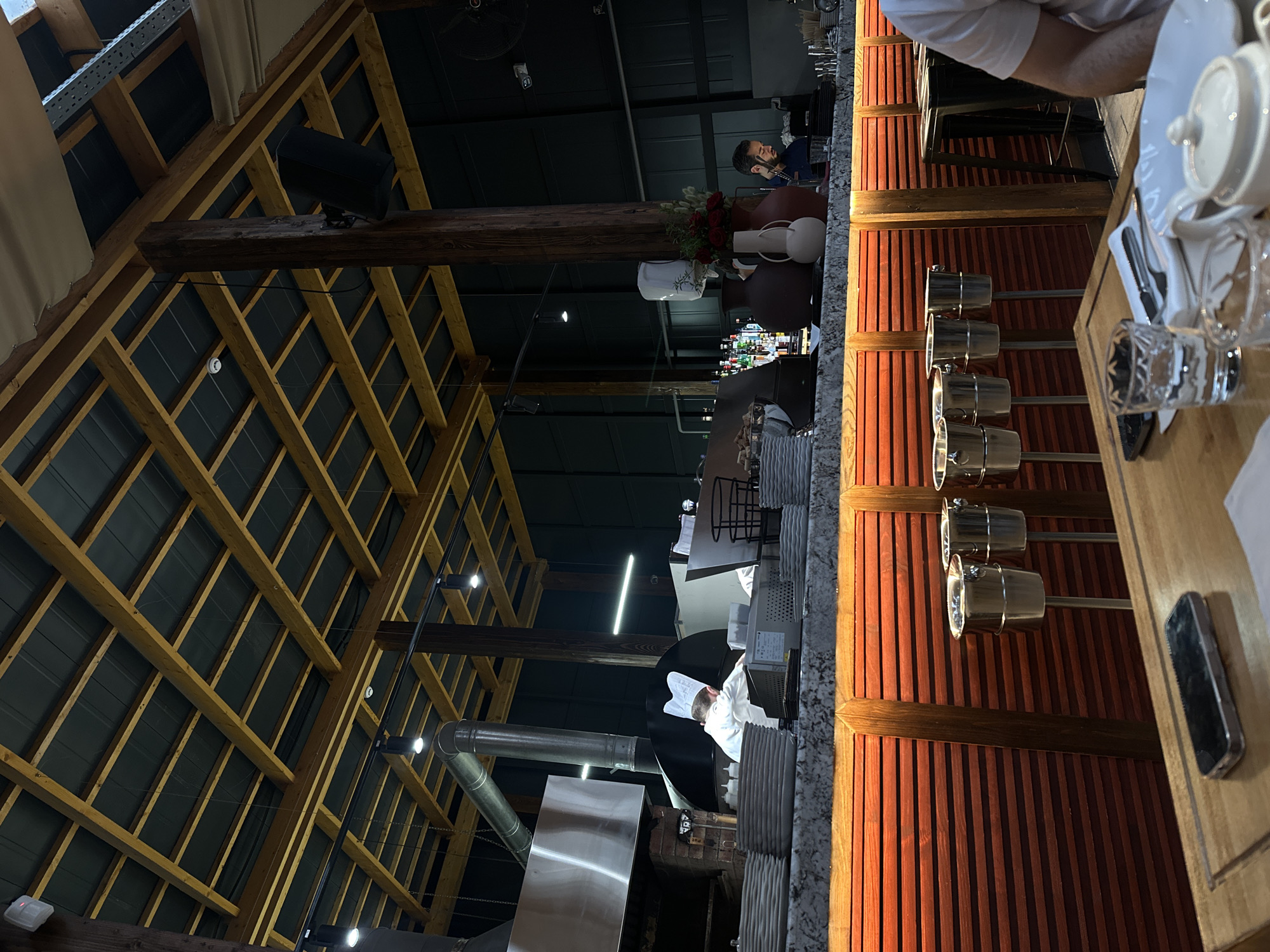 The image presents an interior view of a building with an intricate wooden beam ceiling structure. A significant dark-colored speaker is mounted on one of the beams. Part of a service counter or bar area is visible, featuring wooden paneling and reflective fixtures.
A person wearing a chef's hat and white uniform is seen working at a station that includes stainless steel appliances and a hood.
At the counter, there are tableware items including cups, plates, and cutlery. A man is visible seated at the counter, holding a phone to his ear. There are floral arrangements and decorative vases on the bar top.
Although the location within Yerevan, Armenia cannot be definitively determined from the image, the overall scene suggests a restaurant or hospitality setting.