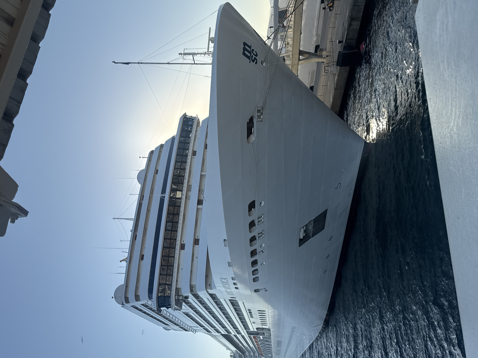 An exterior, eye-level, medium shot captures the bow of the large white cruise ship MSC Musica docked at a port under a clear blue sky. The ship dominates the frame, its hull gleaming in the sunlight. Several rows of windows are visible along its side, as are life rafts. The name "MSC MUSICA" is clearly visible on the ship's superstructure. The ship is moored with thick ropes to the dock. The dark blue water of the harbor is choppy, with sunlight reflecting off its surface. In the background, parts of the port infrastructure, including a dock and a building, are visible. A flag flies from a tall mast on the ship. The scene appears to be set in the port of Ibiza, Spain, given the context of the provided crops which indicate this location. There are no people prominently featured in the image, though the presence of dock structures suggests activity in the port. The overall lighting suggests it is daytime, likely morning or late afternoon due to the angle of the sun.