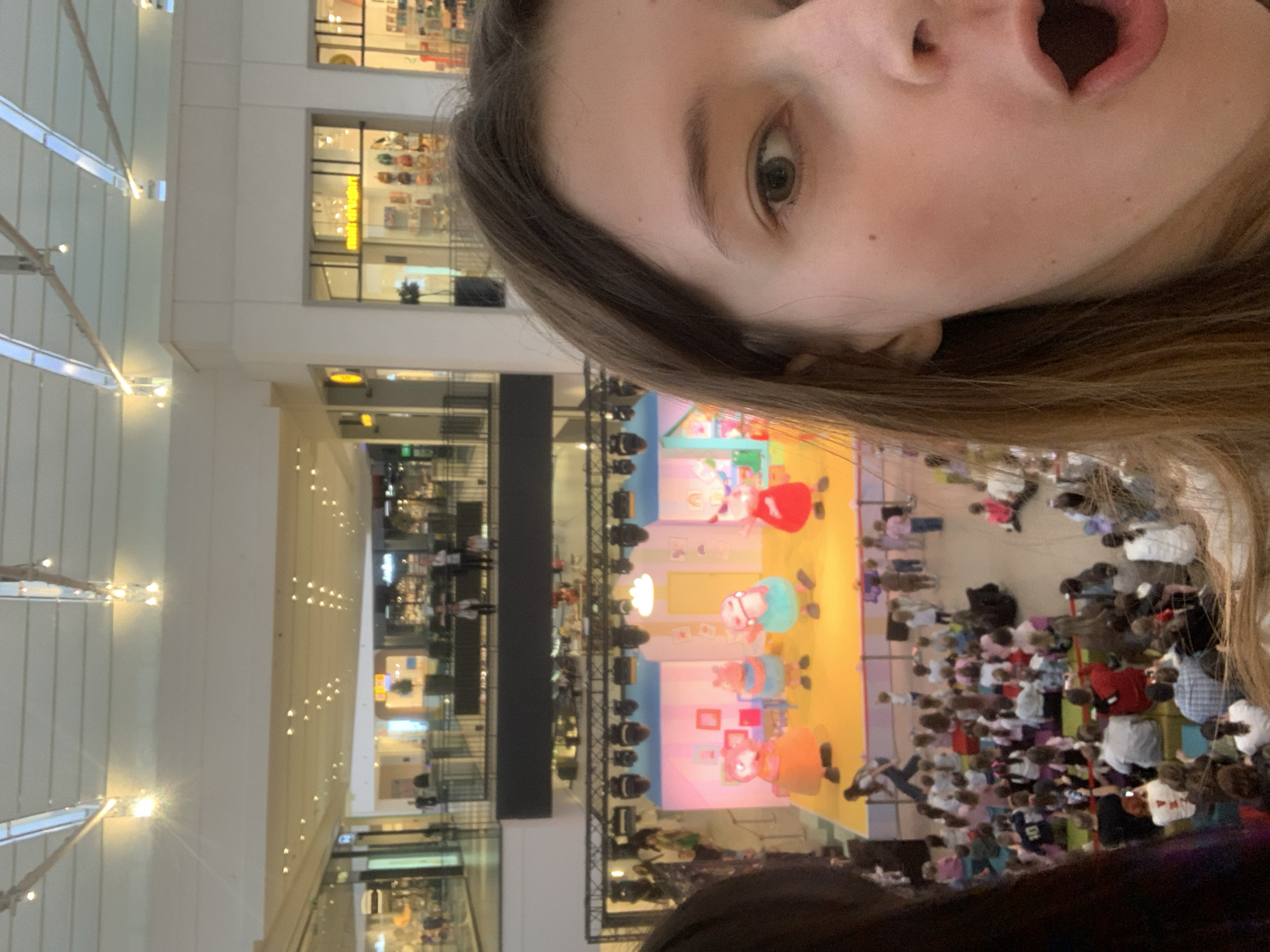 This image captures a first-person perspective of an event taking place inside a multi-story shopping mall in Belgrade, Serbia. The face of an individual, likely the photographer, occupies the foreground, rotated approximately 90 degrees clockwise. The person's left eye is visible, and their mouth is wide open in an expression of surprise or excitement. Long, brown hair frames the side of the face.
In the background, on the ground floor of the mall, a vibrant children's performance is underway on a brightly lit stage. The stage features several large, colorful cartoon character mascots, possibly resembling pigs, suggesting a show like Peppa Pig. A considerable crowd of people, including many children, is gathered in front of the stage, attentively watching the performance.
The setting is an expansive indoor space with multiple levels, featuring white architecture, balconies, and artificial lighting. Shopfronts are visible on the upper floors. A yellow sign in the background shows blurry, indistinct text. The atmosphere appears lively and engaging, consistent with a family-oriented public event within a commercial complex.