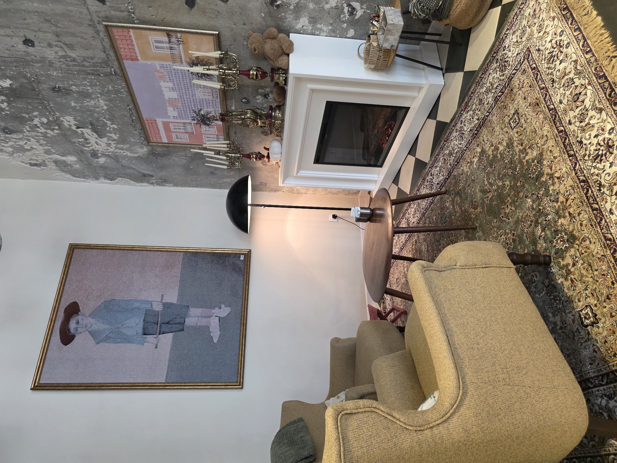 The image depicts an interior scene, possibly a cafe or lounge, with a rustic and eclectic aesthetic. In the foreground, two plush, textured tan armchairs are angled towards the center, one in sharper focus than the other. A dark wood, circular coffee table with three tapered legs sits between them, holding a small metallic container and what appears to be a sugar packet.
Behind the coffee table, a tall, black arc floor lamp with a dome-shaped shade casts a warm glow. Further back, a white mantelpiece fireplace is the focal point of the right side of the room. It is adorned with several decorative items, including a golden clock, ornate candelabras with white candles, a white pumpkin, and a plush brown teddy bear. Above the fireplace, a framed painting depicts colorful buildings, possibly in a European style.
Dominating the left wall is a large, framed portrait of a young boy in a straw hat, wearing a blue jacket and shorts, holding a stick. The walls are a mix of white and rough, textured concrete. The floor is covered by a large, ornate rug with intricate floral patterns in shades of green, beige, and burgundy. The rug lies on top of a black and white checkered floor. To the right of the fireplace, a small side table holds woven baskets filled with various items, suggesting a display or retail area. The overall impression is one of cozy, artistic, and slightly bohemian ambiance. The presence of a faux fireplace and decorative elements suggests a deliberate attempt to create a comfortable and visually engaging space. The image does not provide any explicit clues to pinpoint its location within Khatay Raion, Azerbaijan.