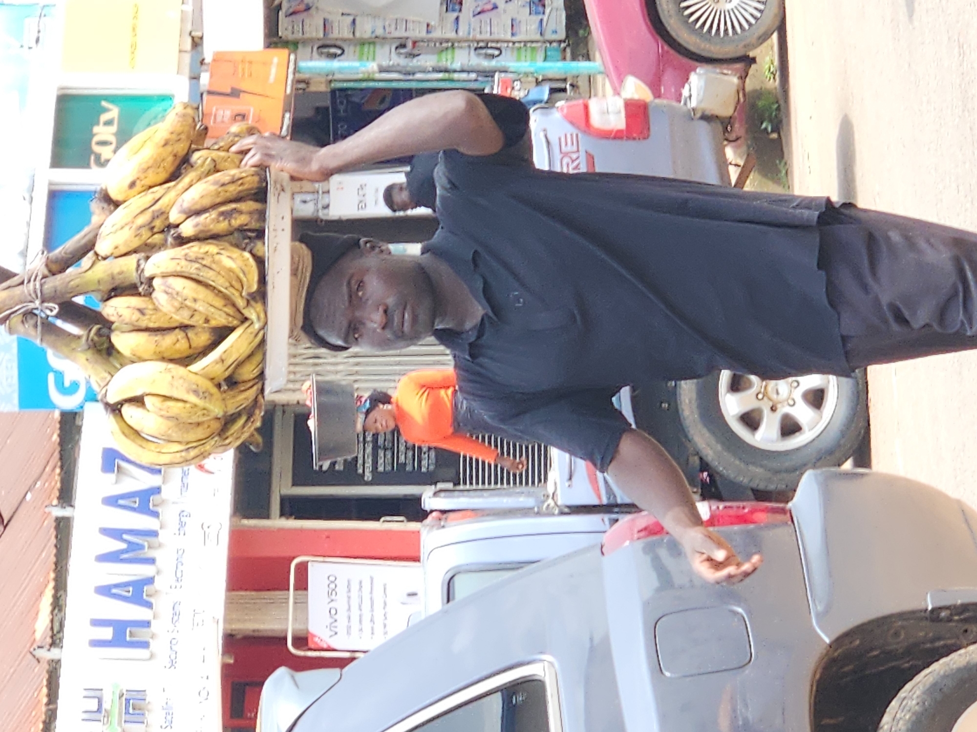 In a street scene in Jos, Nigeria, a dark-skinned man walks with a large bunch of bananas balanced on his head. He is wearing a black polo shirt and black trousers. Behind him, a woman in an orange top and black skirt also carries a basin on her head. The background shows shops with signs, including one that reads "HAMAZ". Cars are visible on the street, including a grey SUV and a red car. The overall environment suggests a bustling market or commercial area.