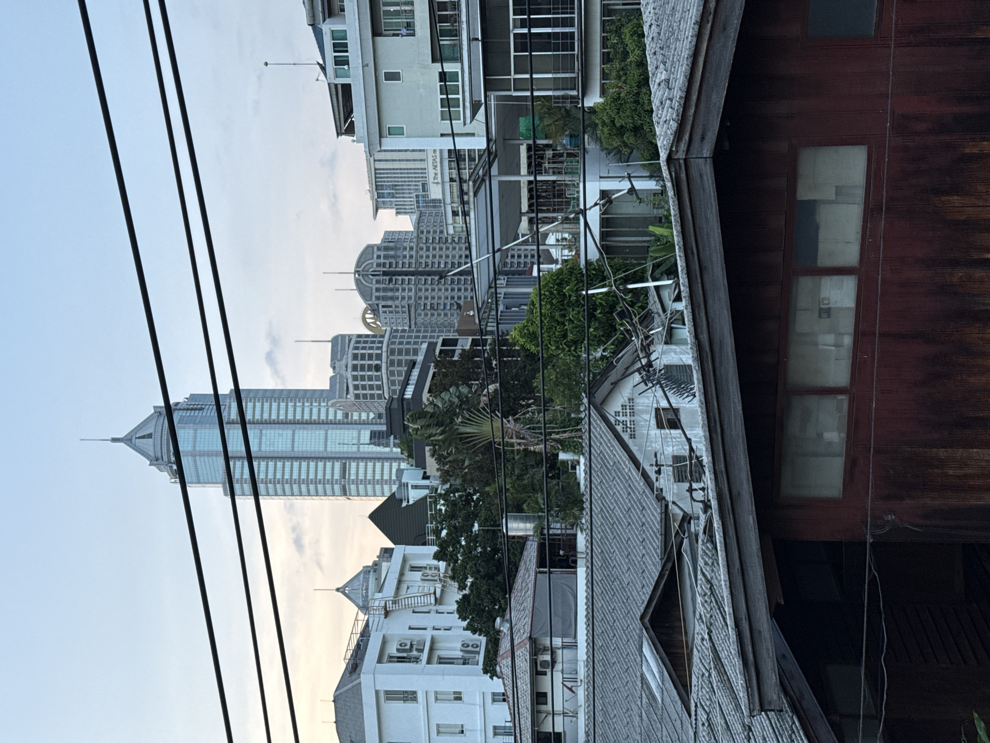 This is a photograph of an urban landscape in Bangkok, Thailand. The image shows a dense collection of buildings, ranging from modern skyscrapers to older, lower-rise structures with tiled roofs. Prominently featured in the background is a tall, slender skyscraper with a pointed spire, reflecting the sky. In the mid-ground, there are several mid-rise buildings with balconies and windows, interspersed with lush green trees. The foreground is dominated by the roofs of older, weathered buildings, one of which has a dark wooden facade and windows. Utility wires are stretched across the upper portion of the image, partially obscuring the sky and some of the buildings. The overall impression is one of a city with a mix of architectural styles and varying degrees of development. The lighting suggests it is either early morning or late afternoon, with a soft, diffused light.