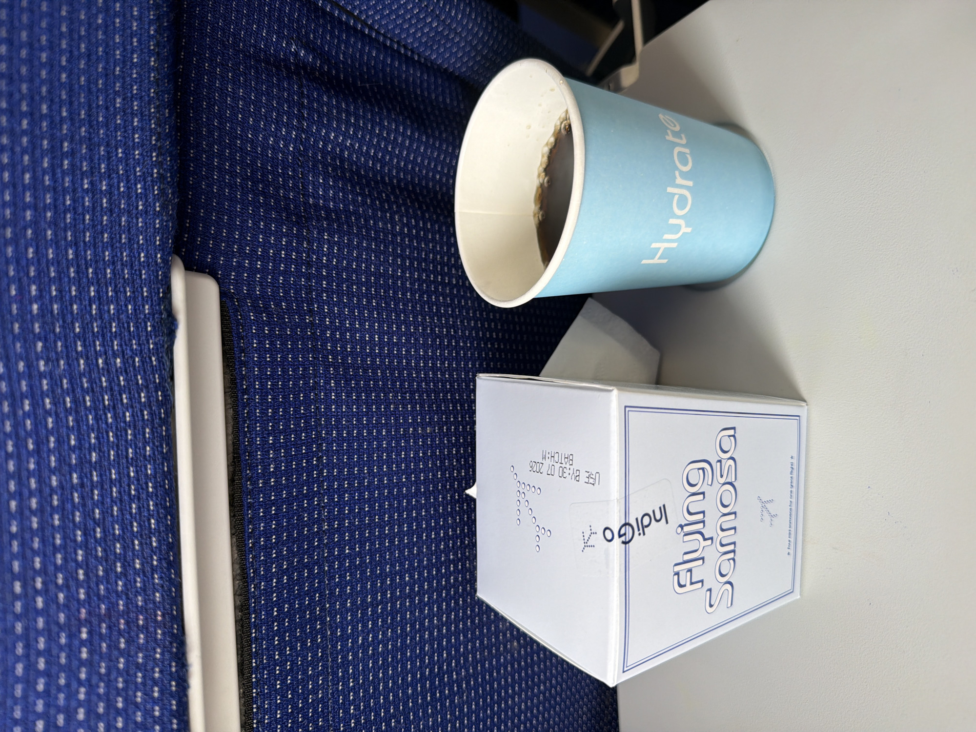 An overhead shot shows a blue Indigo Airlines branded box labeled "Flying Samosa" next to a light blue cup with the word "Hydrate" printed on it, containing a dark beverage. The items are on a light-colored tray table, likely in an airplane cabin, with the blue, patterned upholstery of airplane seats visible in the background. There are no people visible in the image. The scene is a close-up of the tray table, with the focus on the food and drink items. The specific location in Mohan, India, is not discernible from the image.
