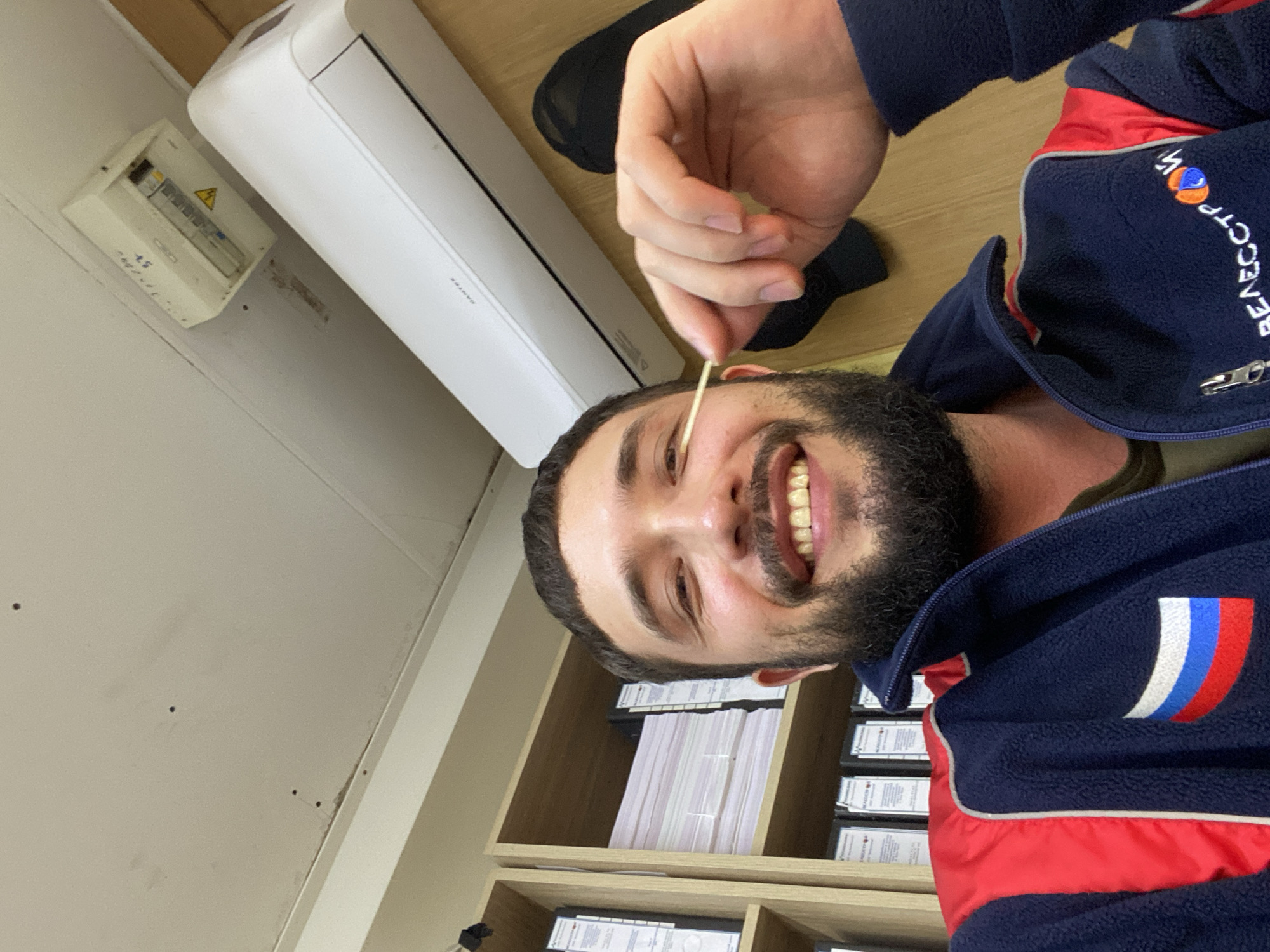 In Luzhitsy, Russia, a man with a beard and a dark fleece jacket adorned with the Russian flag and a company logo is taking a selfie. He is holding a wooden stick to his eye. In the background, shelves are filled with binders and papers, and an air conditioning unit is mounted on the wall near an electrical panel.