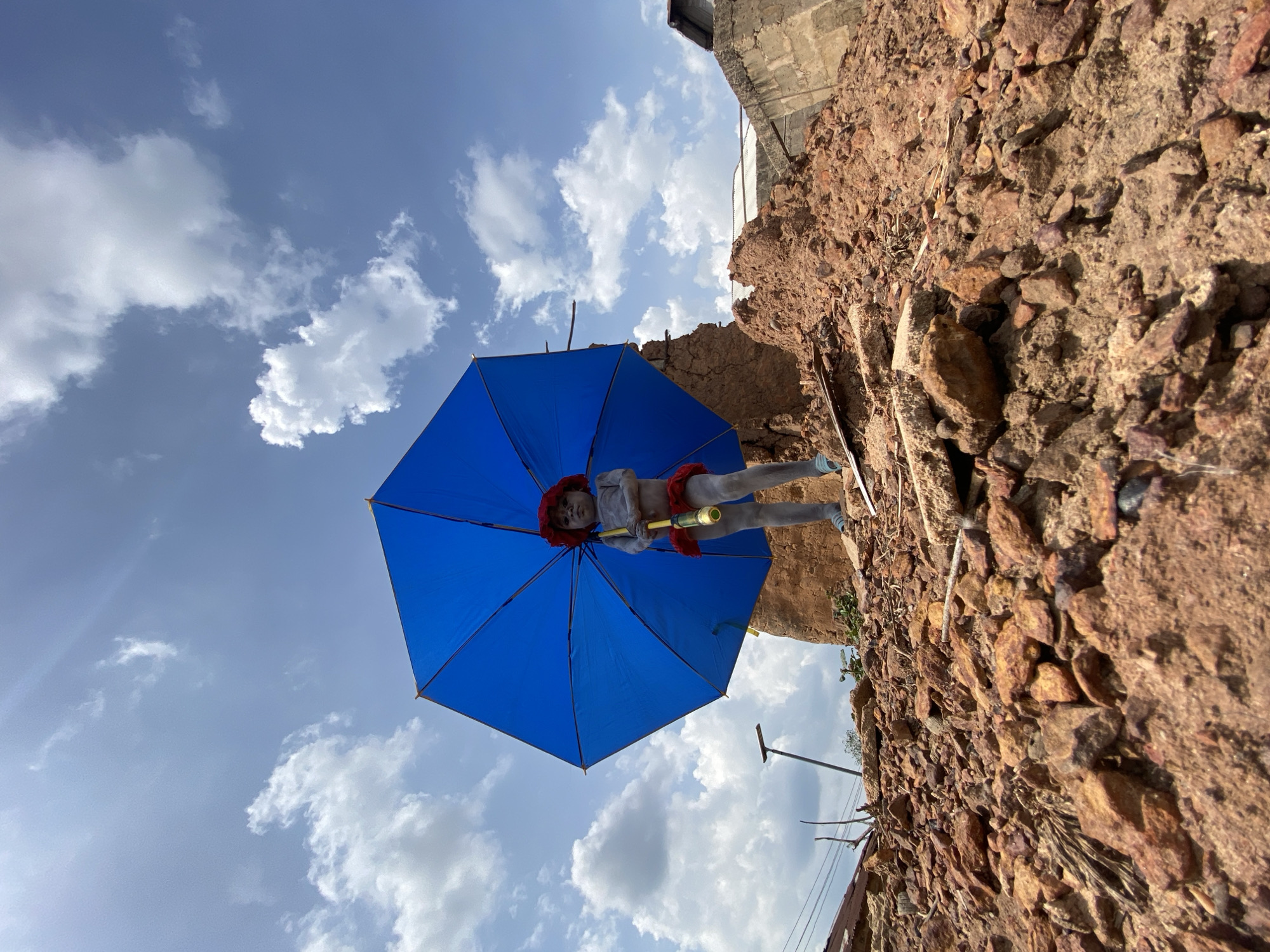 A blue umbrella, held by a nude-colored figurine with red head covering and trim, is seen against a cloudy sky in Amoso, Nigeria. The figurine is holding a yellow and green pole. To the right of the figure is a wall comprised of rocks and debris. A utility pole with wires is also visible.