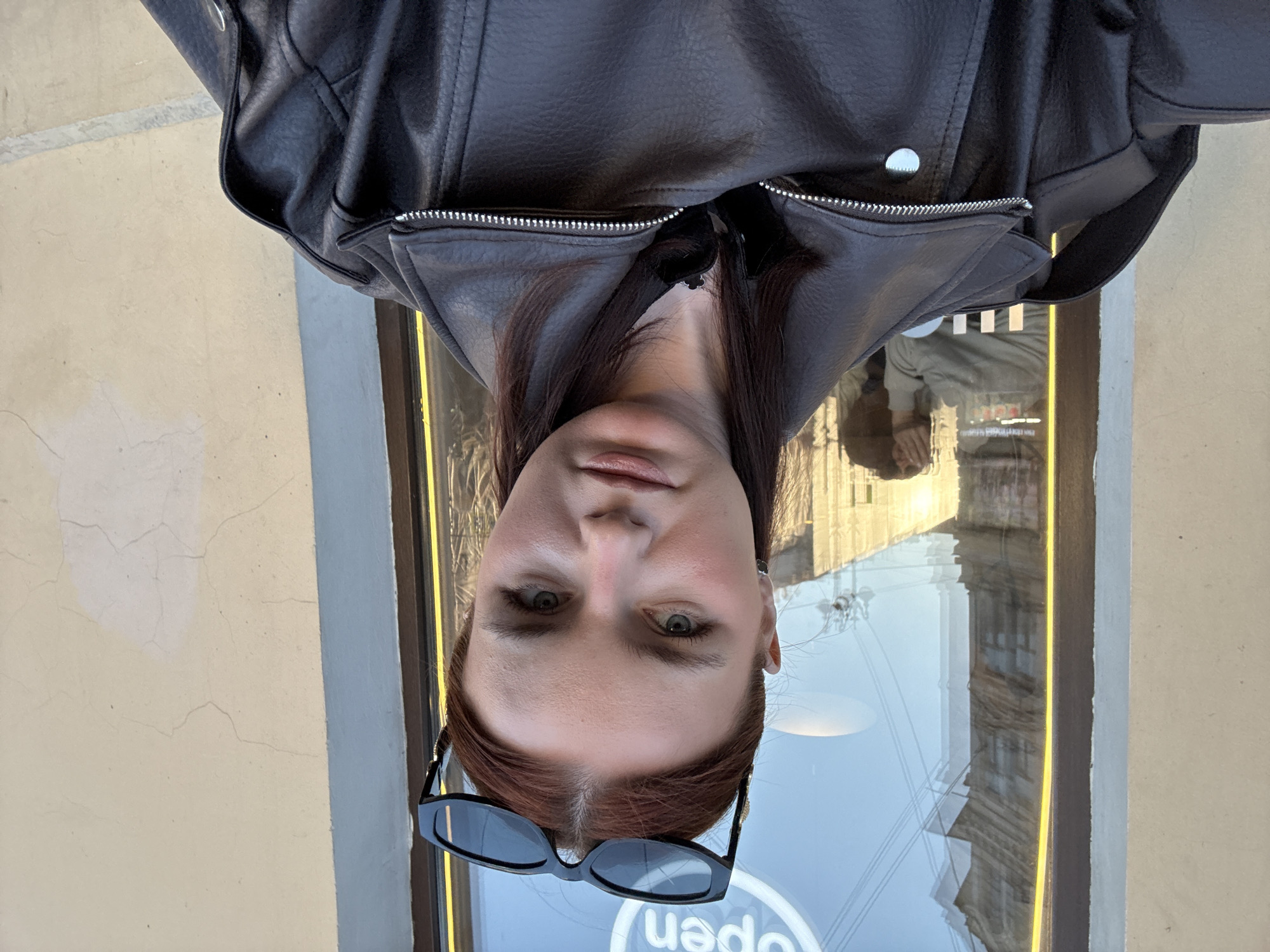 A woman stands in front of a window in Saint Petersburg, Russia, wearing a black leather jacket and sunglasses. The reflection in the window reveals architectural details of the city. The window also shows a sign that reads "Open". The wall next to the window is pale yellow with cracks.