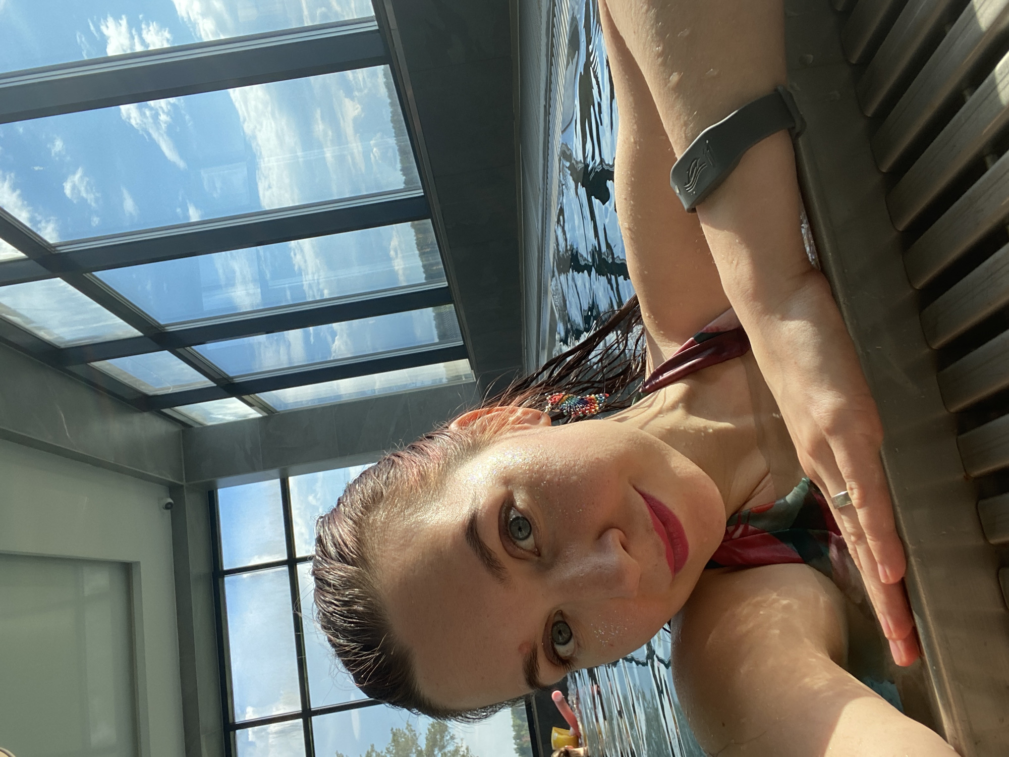 A woman is taking a selfie in an indoor swimming pool. She has wet, reddish-brown hair, blue eyes, and is wearing pink lipstick. She has glitter on her cheeks and is wearing a colorful beaded earring in her right ear. Her left arm is resting on the edge of the pool, which is made of wooden slats and a gray border. She is wearing a ring on her left hand and a dark gray fitness tracker on her right wrist. She is wearing a red and green patterned swimsuit. In the background, large windows show a blue sky with white clouds and trees outside. Another person is visible in the water to the left, holding a pink floatation device. The location is the pool area of the Kama River Resort in Chaikovsky, Russia.