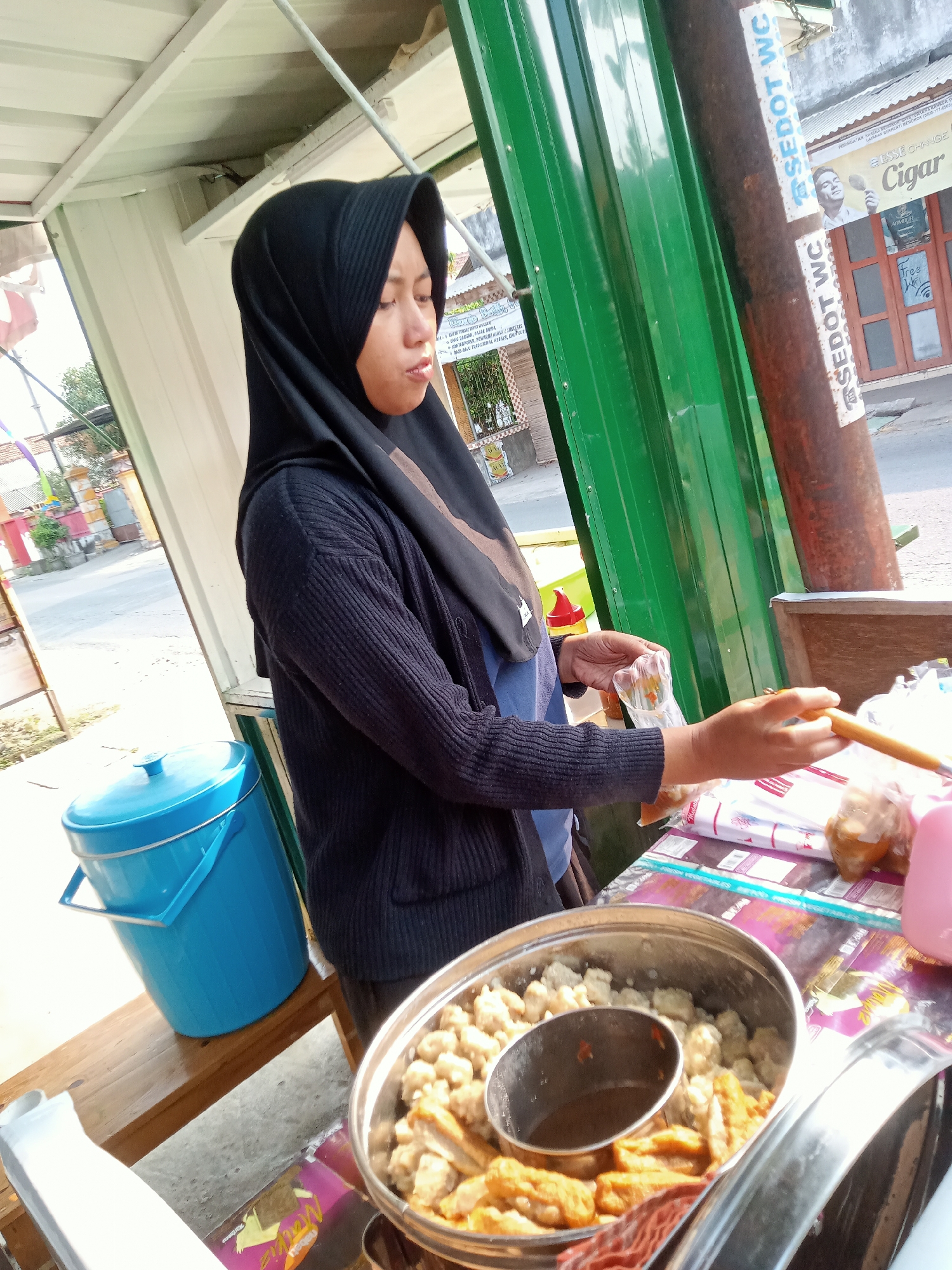 A woman wearing a black hijab and dark blue sweater stands behind a food stall in the city of Simo, Indonesia. She is preparing food, holding a plastic bag in her left hand and a skewer or stick in her right hand. In front of her is a metal bowl containing various fried foods, including meatballs and what appear to be fried tofu or fish cakes, along with a small bowl of dipping sauce. A blue insulated cooler is visible to her left, and a wooden bench is positioned beneath it. The stall has a green metal structure and a white corrugated metal roof. In the background, a pole with stickers that read "SEDOT WC" is visible, along with storefronts advertising "Cigar" and "Free Wifi." The overall scene depicts a street food vendor at work in an urban environment.