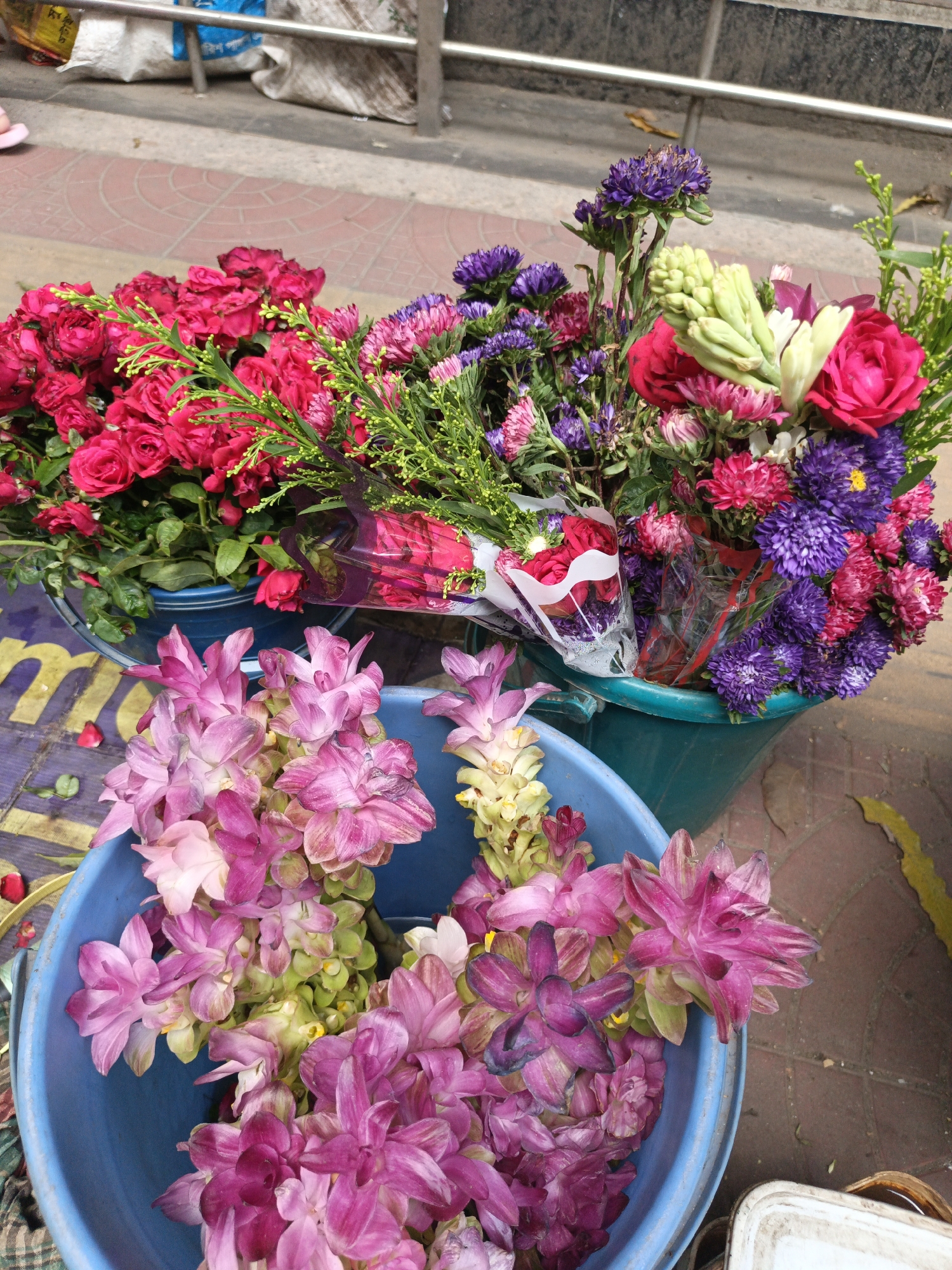 The image shows three buckets of flowers for sale on a street in Dhaka, Bangladesh. In the foreground, a blue bucket is filled with pink and purple Curcuma flowers. Behind it, a green bucket contains a mix of red roses, purple asters, and white tuberose. Another green bucket on the right also holds various flowers. The buckets sit on a sidewalk with red brick paving and are partially obscured by bags and a metal railing in the background. The environment is urban and suggests a street vendor or florist stall.