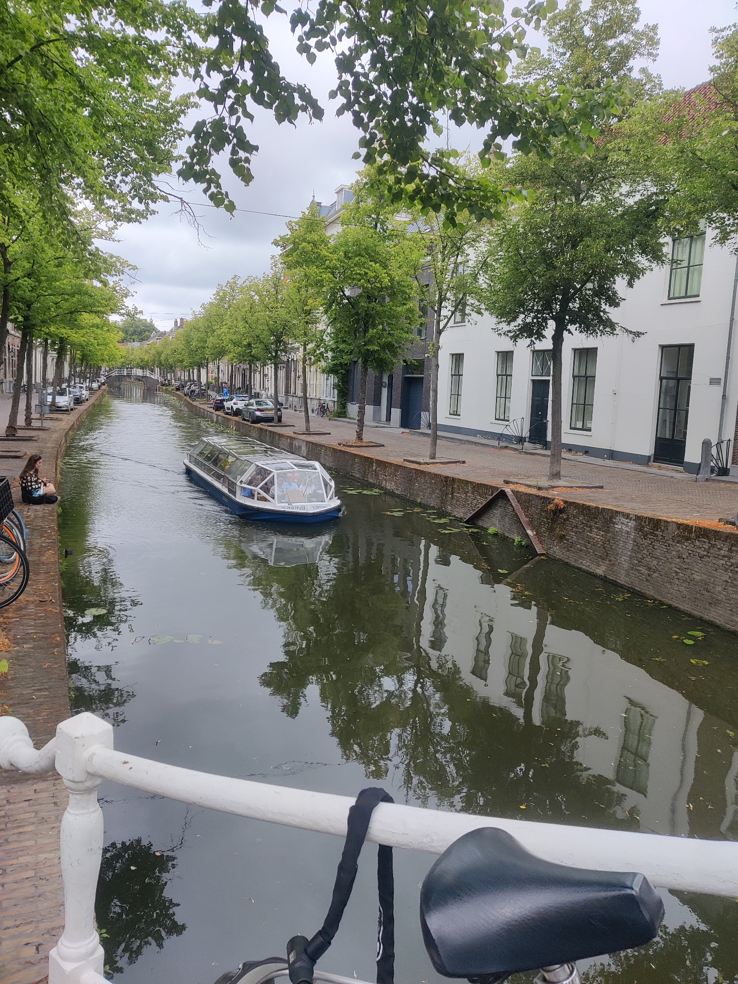 The image depicts a canal in the city of Delft, Netherlands, likely in the historic city center, given the presence of traditional Dutch architecture. The canal is lined with trees and multi-story buildings. A glass-enclosed tour boat is navigating the canal. On the left bank, a young woman is sitting on the brick embankment, facing the water, and looking at her phone. A bicycle is parked near her. The water reflects the surrounding buildings and trees. The sky is overcast. The perspective is from a bridge, looking down and across the canal.