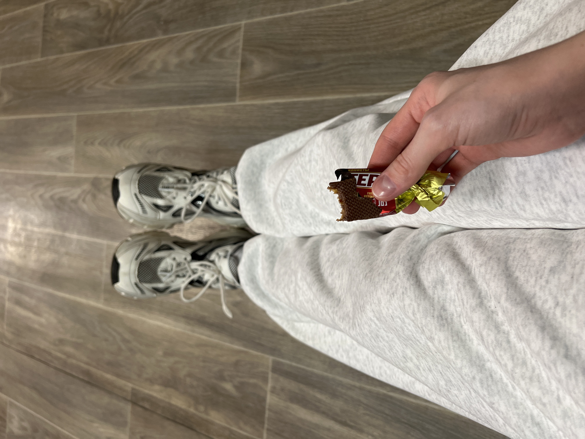 An overhead shot shows a person sitting on a wooden floor in a casual outfit consisting of gray sweatpants and white sneakers with gray and black accents. The person is holding a Стеp (Step) chocolate bar in their right hand, from which they have taken a bite. The chocolate bar is partially unwrapped, with the gold wrapper visible. The location is assumed to be Khimki, Russia, based on the Cyrillic characters on the chocolate bar packaging, which suggest a Russian origin.