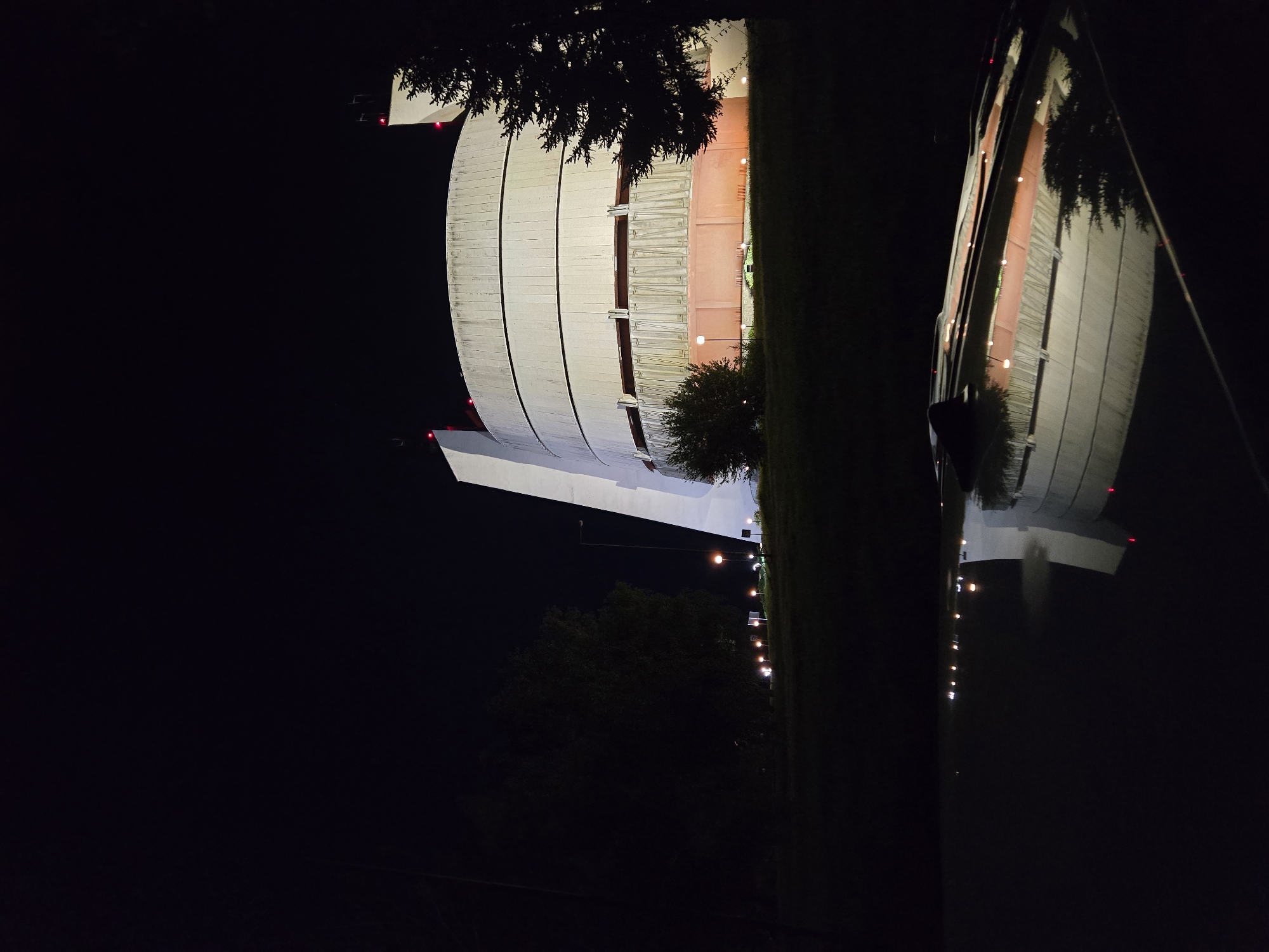 The image depicts the Pleven Panorama museum in Pleven, Bulgaria at night. The museum's distinct cylindrical structure is illuminated, contrasting against the dark sky. A tree and vegetation are visible in the foreground. Streetlights line the horizon to the right of the museum. The image is oriented sideways.