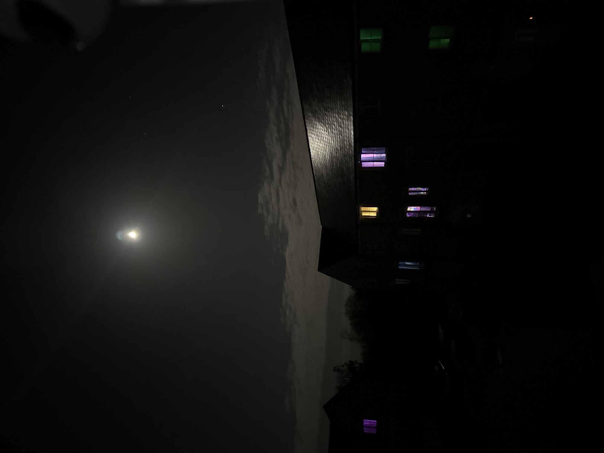 This image depicts a nighttime exterior scene in Great Yarmouth, United Kingdom. The sky is dark with notable cloud cover, illuminated by a prominent, bright moon positioned in the upper-center, casting a distinct halo and slight reflections on the building roofs. Several faint stars are visible in the upper sky.
In the midground, a multi-story building, likely residential, dominates the right half of the frame. It features a dark grey tiled or slated roof that reflects ambient moonlight. Several windows on this building are illuminated: an upper-floor window on the far left displays a yellow-orange light; an adjacent upper-floor window to its right emits a strong purple light; and an upper-floor window on the far right shows a green light. On a lower floor, a window to the left displays multiple small, colorful lights, while another window further right on the same floor shows a bluish-white illumination. The building's facade is otherwise dark.
To the far left, a second, partially obscured building silhouette is visible, behind which dark tree lines are discernible. One window on this left-hand building displays a magenta-purple light. The area between the viewer and the buildings, constituting the foreground and ground level, is profoundly dark, with no discernible features or activities. No individuals are visible anywhere in the scene. An indistinct dark object is partially visible in the upper right corner, likely part of the camera or an immediate foreground obstruction.