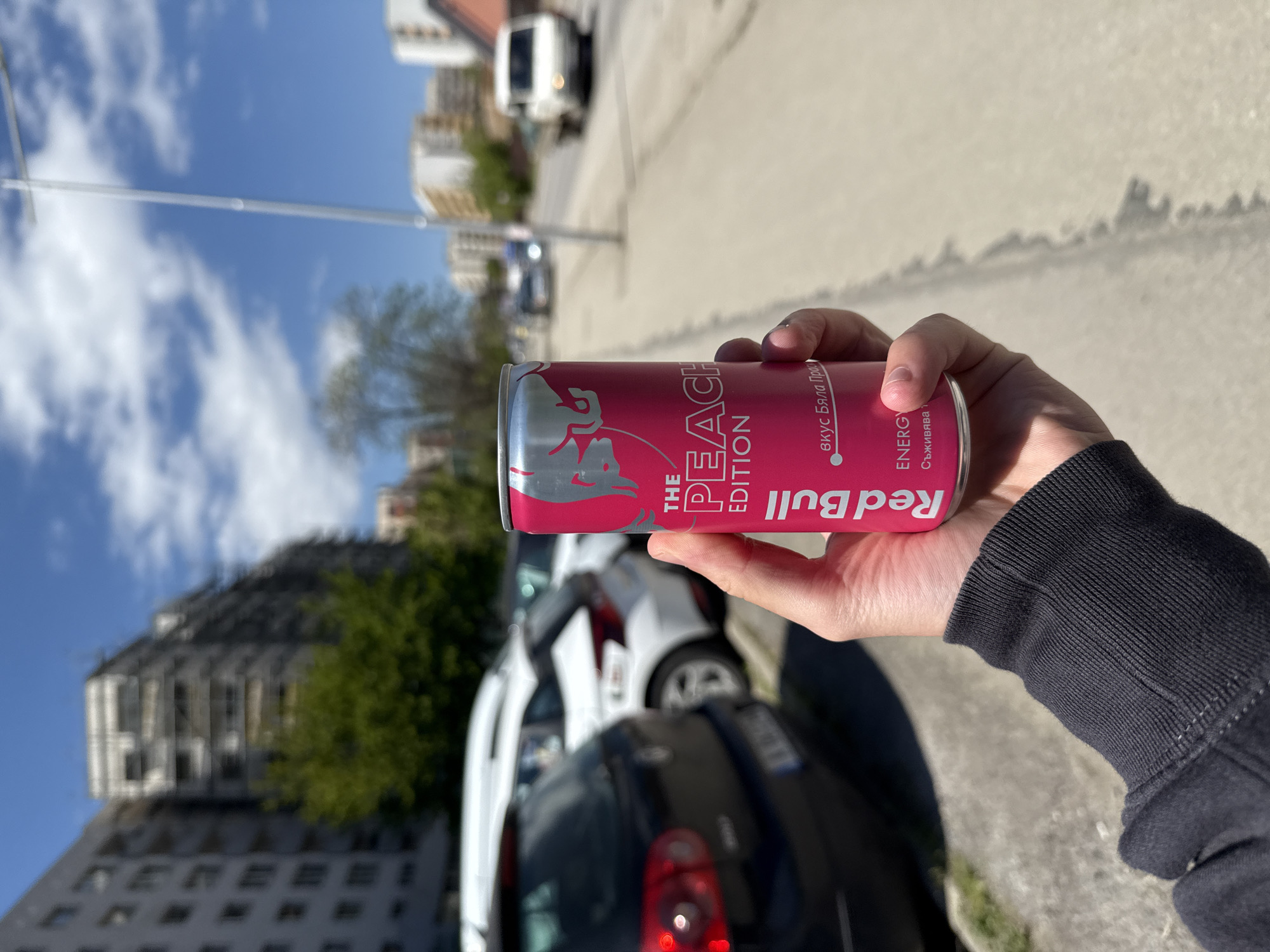A person is holding a can of Red Bull The Peach Edition in front of an urban scene. The can is primarily pink and silver. The person is wearing a dark long-sleeved garment. The background features a street with parked cars (black and white) and buildings. A tall, grey building stands in the distance. The sky is blue with scattered clouds. The text on the Red Bull can appears to be in Bulgarian, indicating a possible location in Bulgaria. Given the architecture and the text on the can, the location is plausibly in the city of Sofia, Bulgaria.