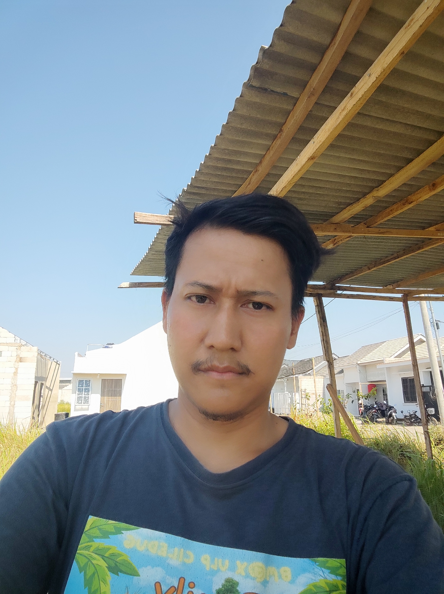 A man is taking a selfie outdoors, possibly in Gebang, Indonesia. He is wearing a dark blue t-shirt with a colorful graphic on the front, and has short black hair and a mustache. Behind him, there is a corrugated metal roof supported by wooden beams, and a bright blue sky. In the background, there are houses under construction and some mature vegetation. The scene suggests an outdoor, possibly semi-rural, environment with ongoing building activity.