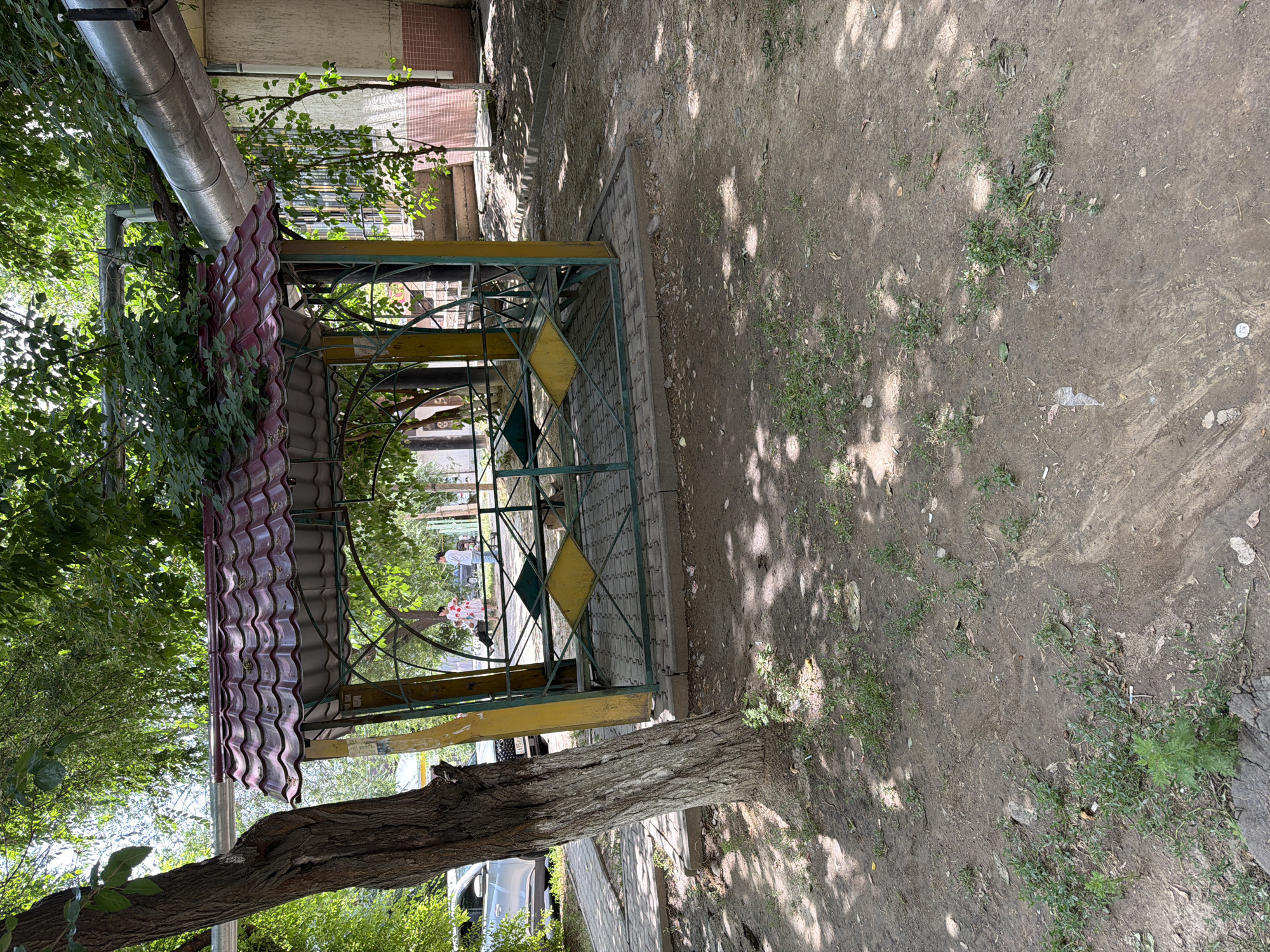 The image depicts an outdoor scene in Shymkent, Kazakhstan, featuring a small, open-air pavilion with a red, tiled roof and yellow and green metal supports. The pavilion is situated on a dirt and patchy grass area, surrounded by trees. In the background, a building with a textured facade and a large, silver pipe overhead are visible. Two individuals, a woman in a patterned dress and a person in a light-colored shirt, are standing inside the pavilion. A silver car is parked to the left of the pavilion. The ground is uneven and shows signs of wear and tear, with scattered debris and small plants growing through the soil. The lighting suggests it is daytime, with dappled sunlight filtering through the trees.