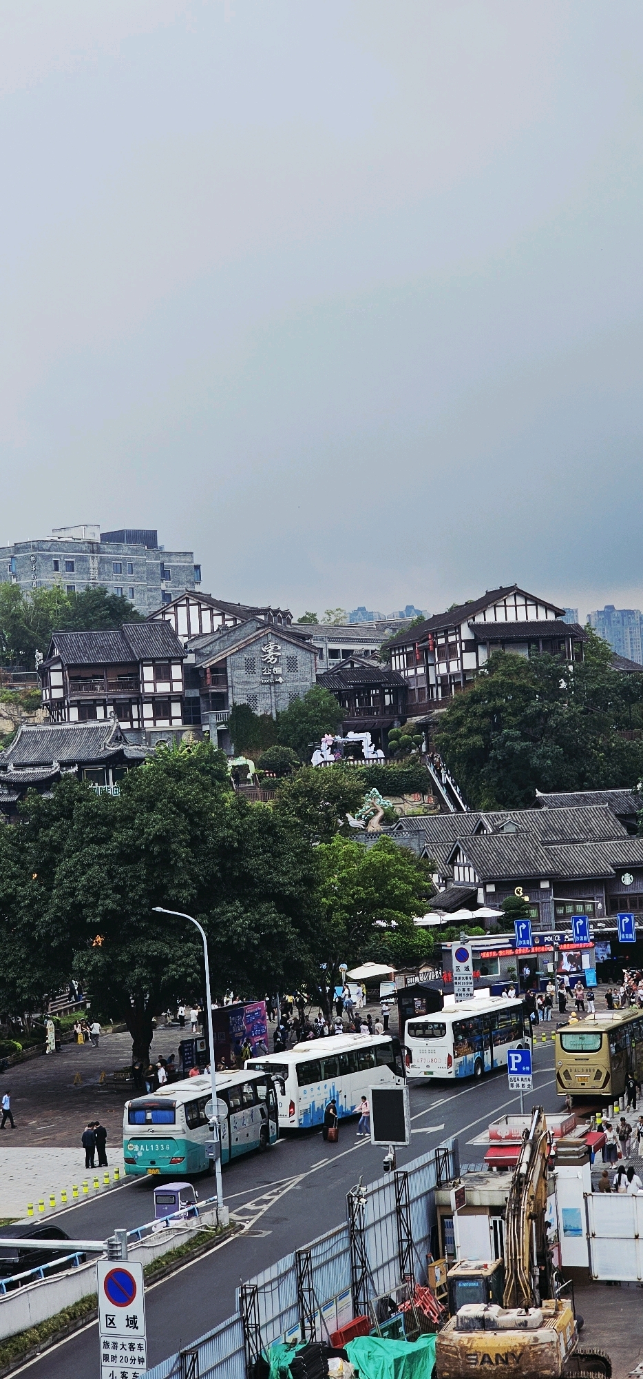 The image captures an overhead view of a street scene in Shapingba District, China. Traditional Chinese architecture with grey tiled roofs and white-trimmed wooden structures dominates the midground, contrasted by a modern grey building in the background. Several buses are parked or driving on the road, including a teal and white bus with license plate AL1336, and a white bus with blue designs. A yellow bus with license plate AL3998 is also visible. A construction site with an excavator and green tarps is in the foreground, behind a metal fence. Pedestrians are present on the sidewalks and near the buses. The sky is overcast, suggesting a cloudy day. Signage in Chinese characters is visible on buildings and street signs.