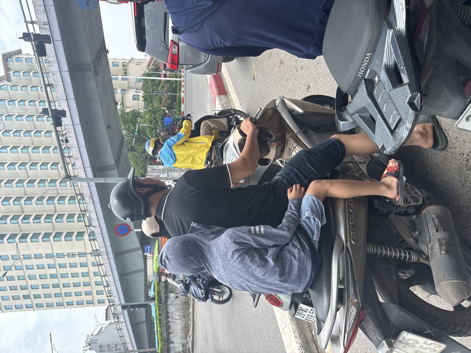 The image captures a street scene in Hanoi, Vietnam, featuring multiple motorcycles and a car amidst urban infrastructure. In the foreground, a child wearing a grey hooded sweatshirt and blue shorts sits on the back of a motorcycle, facing away from the camera. To their right, a person in a black shirt and helmet is seated at the front of the same motorcycle, holding the handlebars. A second motorcycle is visible to the left, with a person in a blue jacket and yellow helmet riding it. Further back, a silver car is stopped, and another motorcycle is partially visible on the right. The background reveals a multi-story building and an elevated road or bridge. The overall environment suggests a busy city street, likely during daylight.
