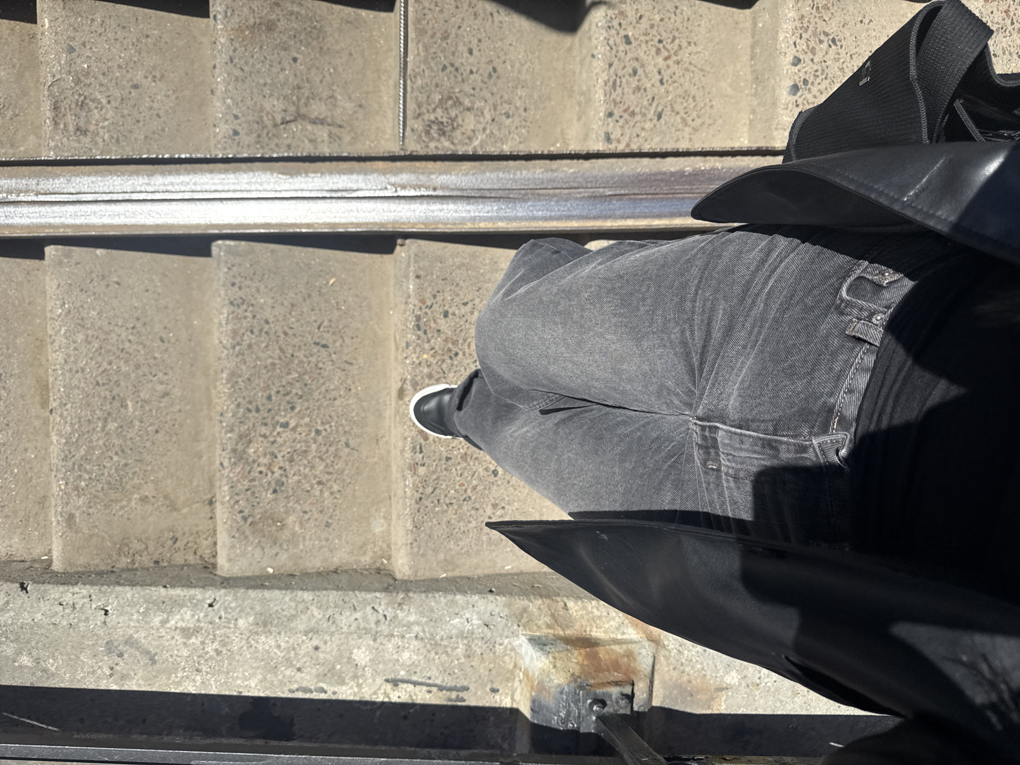 An eye-level, overhead shot depicts a person on concrete stairs near tram tracks. The person is wearing faded black jeans and black shoes with white trim, and a long black leather jacket. The surrounding environment is characterized by weathered concrete steps and tram rails. The location is Moscow, Russia.