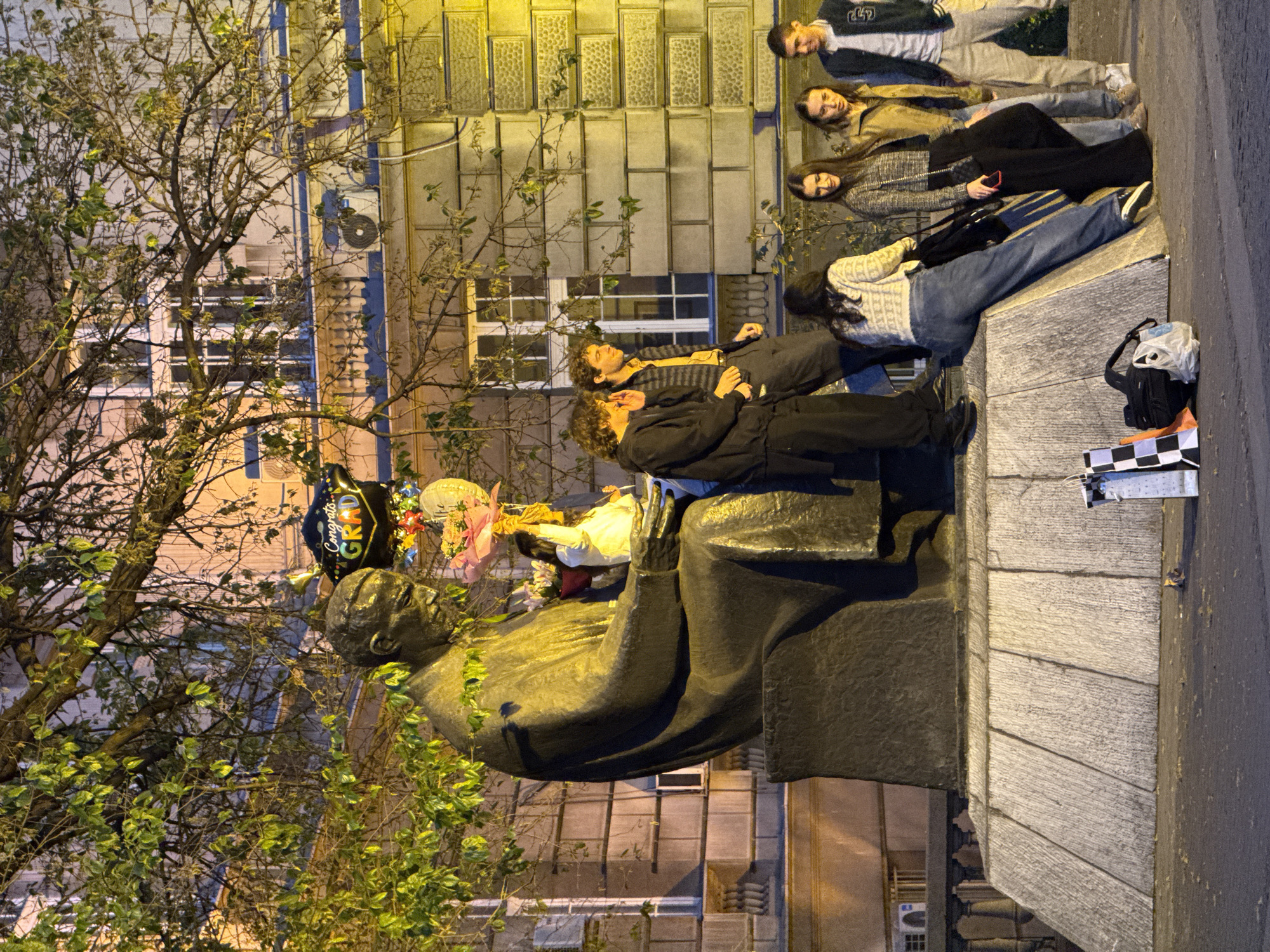 This night-time photograph, taken in Belgrade, Serbia, captures a gathering of young people around a large bronze statue. On the left, the statue depicts a seated man in formal attire, possibly a historical figure. Near the statue's head, a black balloon prominently displays the text "Congrats GRAD!", accompanied by colorful floral decorations, indicating a graduation celebration.
On the right side of the image, a group of approximately eight casually dressed young individuals is present. Some are standing, while others are seated on a low stone base or ledge, suggesting a relaxed, social gathering. Two young men are positioned closer to the statue, one seated and looking upwards, the other standing behind him. Further to the right, several young women and men are standing or sitting, engaged in conversation or observing their surroundings.
The background features illuminated buildings, visible through the sparse branches of a large tree that occupies the left foreground. Artificial lighting from streetlights and building interiors casts a warm glow across the scene. On the ground next to the group, a black backpack, a white plastic bag, and a box with a black and white checkered pattern are visible. The overall mood appears celebratory and convivial.