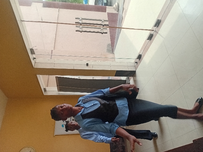 The image portrays an indoor scene, likely a building lobby or hallway, in the city of Ganme, Nigeria. Two African women are in motion, walking from left to right. The foreground subject is an adult female with dark skin and braided hair, wearing a light blue collared shirt, a dark vest, a dark pencil skirt, and dark flat shoes. She carries a dark shoulder bag on her left side and has her right arm extended forward, looking directly at the camera. Behind her, a second female, partially obscured, wears a similar light blue collared shirt and dark trousers, with her hair styled in two dark puffs. She is looking slightly to the left of the camera. The interior features light mustard yellow walls and a floor tiled with light beige square tiles. A round white wall clock, displaying approximately 10:50, is mounted on the upper left wall, above a white banner with a partial blue logo. On the right side of the frame, large transparent glass doors with vertical silver handles offer a view to the exterior, revealing a light brown building facade, pavement, and green foliage in the background under daylight. A dark, open doorway is visible on the far left of the image.