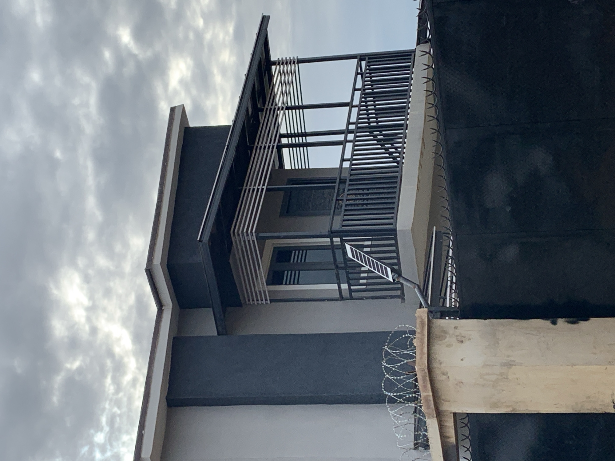 The image depicts a partial exterior view of a modern two-story building in Rafin Par, Nigeria, set against an overcast sky. The building features light grey and charcoal grey walls, with a flat roofline. A prominent second-story balcony is present, enclosed by a dark metal railing with vertical balusters. The balcony is covered by a dark metal awning supported by vertical beams, with horizontal decorative or shading slats positioned both above the railing and below the awning. A dark-framed double door or large window with horizontal elements is visible behind the balcony railing. A white rectangular solar panel light fixture is mounted at an angle on the balcony railing, connected to a dark downward-sloping pipe. In the lower left foreground, a light-colored concrete pillar is topped with multiple coils of razor wire. A dark, textured metal gate with sharp security spikes along its top edge is partially visible behind the pillar, extending towards the right. No people are visible in the scene.