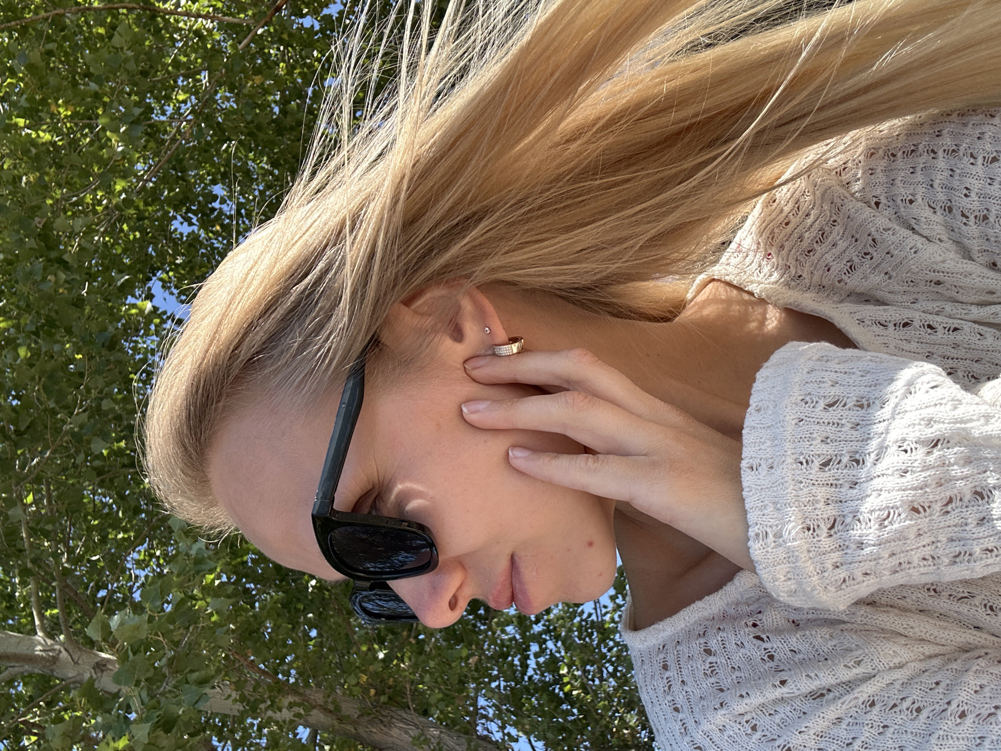 A fair-skinned blonde woman with long, wavy blonde hair and fair skin is captured in a close-up profile shot. She is wearing black sunglasses and a white, open-knit sweater. Her right hand is touching her cheek, with her fingers extended. The background is a blur of green foliage from trees, suggesting an outdoor setting. The lighting indicates it is daytime, with sunlight filtering through the leaves, creating dappled patterns on her skin and clothing. The location is stated to be the city of Gornyi Erik, Russia.
