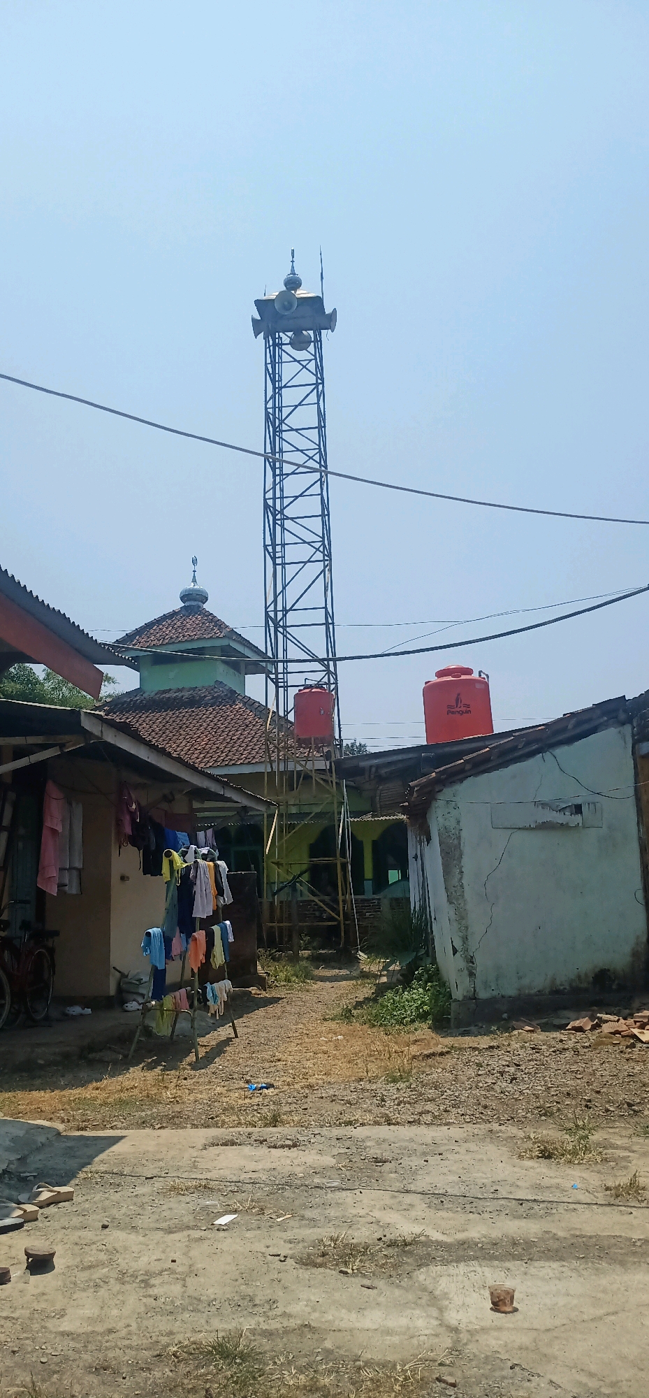The image shows a scene in Bajong, Indonesia, on a bright, clear day. Dominating the center of the frame is a tall, black metal lattice tower equipped with multiple speakers at its apex, likely for public announcements or prayer calls. To the left of the tower is a mosque with a green dome and a tiled roof. In front of the mosque, clothes are hanging on a drying rack. Adjacent to the mosque is a simple house with laundry hanging outside. Two bright orange water tanks, one labeled "Penguin," are visible on rooftops near the tower. The ground is covered with a mixture of dry earth, gravel, and some sparse vegetation. The overall environment appears to be a residential area with a mix of religious and domestic structures.