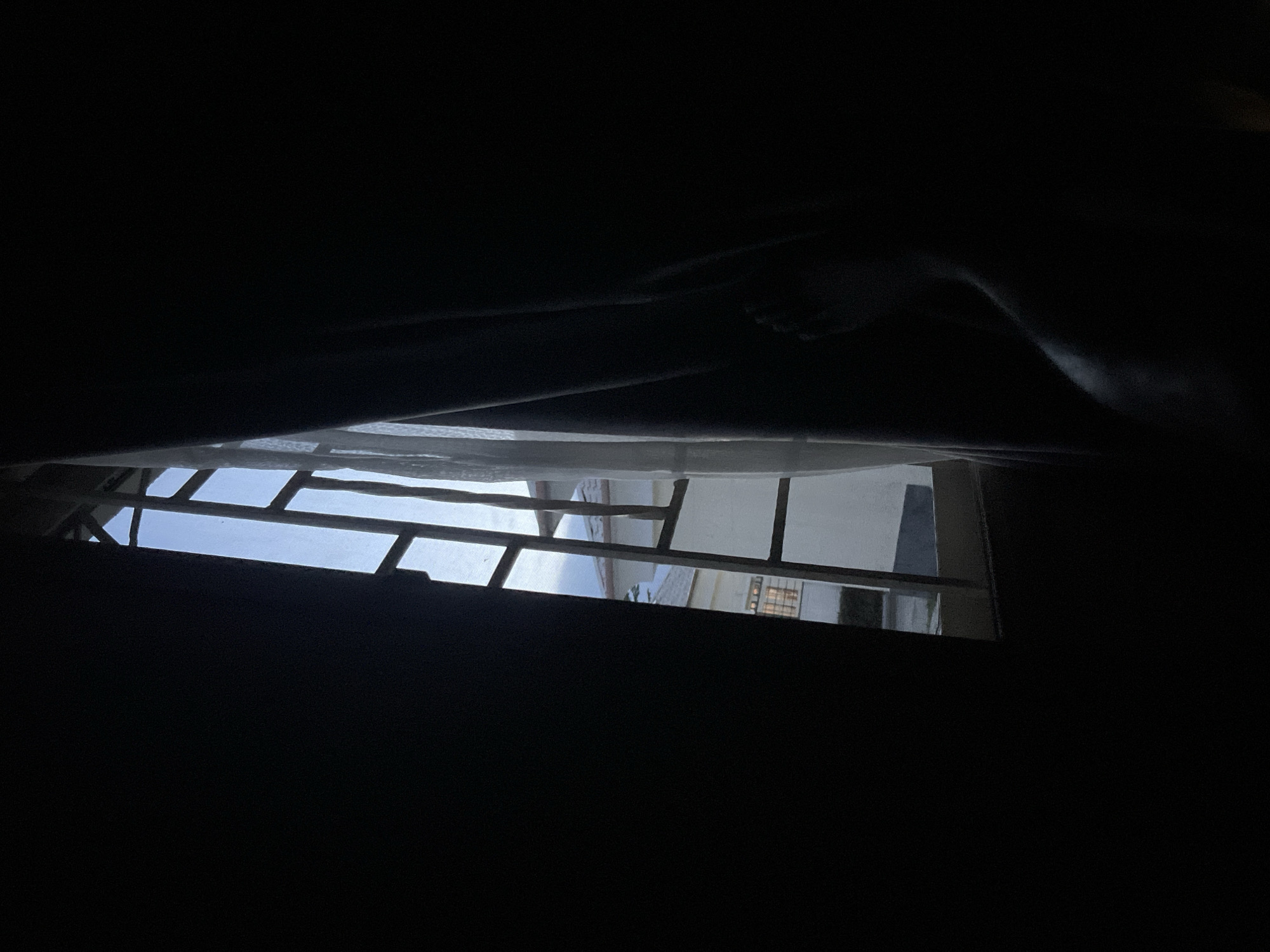 The image appears to be taken from inside a dark room, looking out through a barred window. The sky and a building exterior are visible through the window. A leg is visible in the top portion of the frame, partially obscured by the darkness. The location is specified as Abuja, Nigeria.