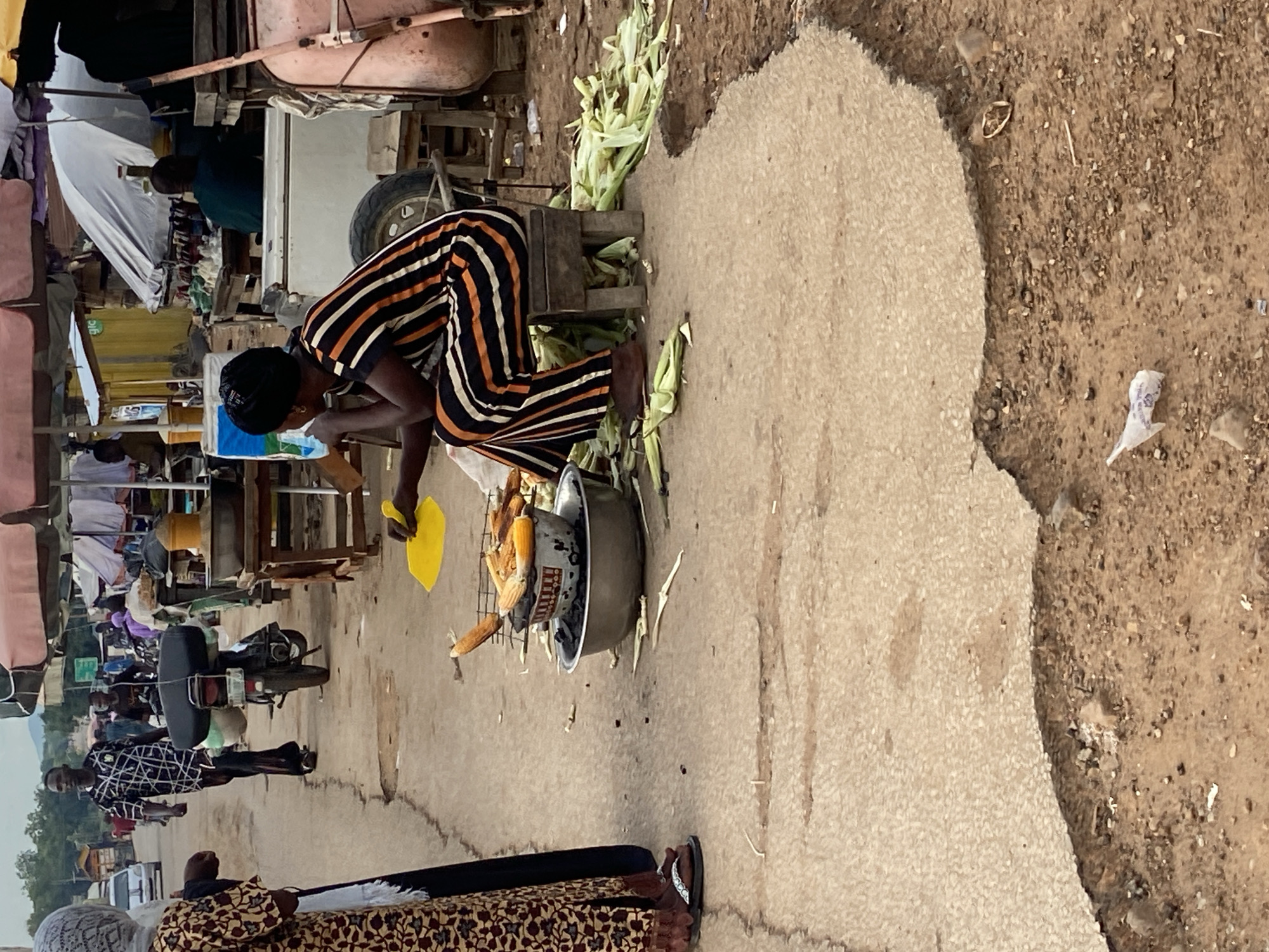 The image depicts an outdoor market scene in Mararaba, Nigeria, observed during daylight hours under an overcast sky. In the immediate foreground, a woman is seated on a low wooden stool on the dirt ground, facing left. She wears a black head covering and a black, white, and orange striped jumpsuit. Her right hand holds a yellow plastic spatula-like object, while her left hand is near her chin. Directly in front of her, several ears of corn are roasting on a metal grate positioned over charcoal within a large metal bowl, which rests inside a wider metal basin. A pile of discarded corn husks and cobs lies on the ground next to her stool. To the left of this setup, a woman is partially visible, standing on a cracked concrete surface. She wears a patterned brown and maroon long dress over dark trousers and flip-flops.
In the midground, a man in a black and white patterned shirt and dark trousers walks on the concrete road, heading away from the viewer. Behind him, a black motorcycle, laden with luggage on its seat, is positioned on the road. The background reveals a lively market environment characterized by numerous stalls, many shaded by large umbrellas of various colors including reddish-brown, white, and yellow. Visible elements within the market include a white cooler, tables displaying items such as bottled beverages, and yellow corrugated metal structures. Other indistinct figures populate the market area. Further down the road on the left, a white car is visible. The ground consists of a worn, broken concrete road that transitions into a dirt shoulder on the right, where small pieces of debris, including a white plastic bag, are scattered. Distant green hills and vegetation are discernible beneath the sky.