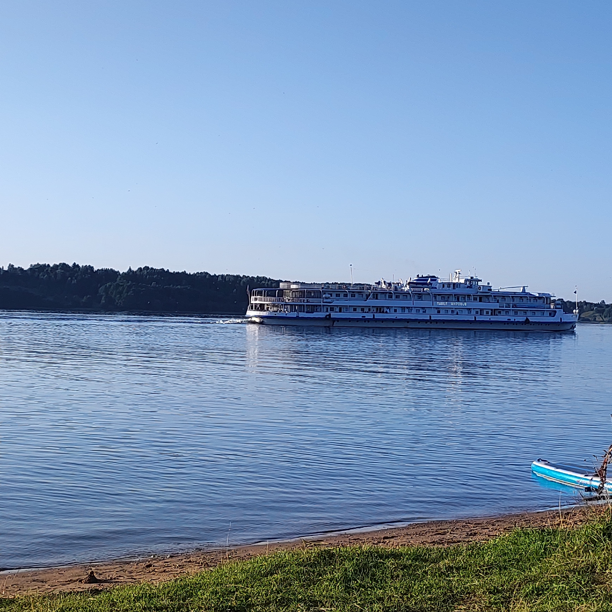 A large white river cruise ship with blue trim is sailing on a wide, calm river under a clear blue sky. The ship has multiple decks with many windows. The shore on the far side of the river is lined with a dense, dark green forest. In the foreground, a sandy beach transitions to green grass. A blue and white inflatable paddleboard rests near the shore on the right side of the image. The scene appears to be set in Navoloki, Russia, given the characteristic landscape of the region.