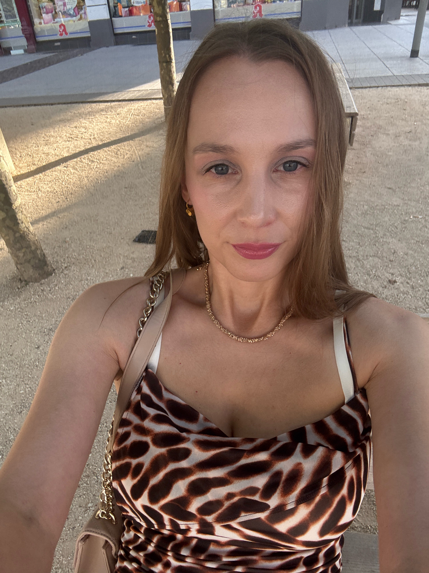 A woman is taking a selfie outdoors in Saarlouis, Germany, likely in the late afternoon due to the warm lighting and shadows. She has fair skin, blue eyes, and light brown hair, and is wearing a leopard-print dress and a gold necklace. She is also carrying a tan purse. The background shows a paved area with benches and trees, suggesting a public space like a park or pedestrian walkway. Buildings with storefronts are visible in the distance, hinting at an urban setting within Saarlouis.