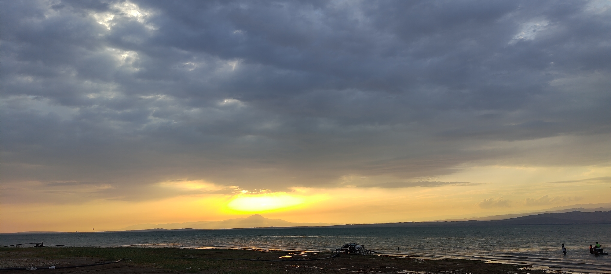 A wide shot captures the sunset over a large body of water, likely a lake or sea, with mountains visible in the distance. The sky is filled with dramatic clouds, with a bright patch of sunlight breaking through above the horizon, casting a warm yellow and orange glow. In the foreground, a dark shoreline is visible, with some debris and equipment scattered on it. To the right, a small boat with two people in it is visible on the water. The overall atmosphere is one of a peaceful evening at the end of the day. The image is believed to have been taken in the city of Chigdalik, Tajikistan.