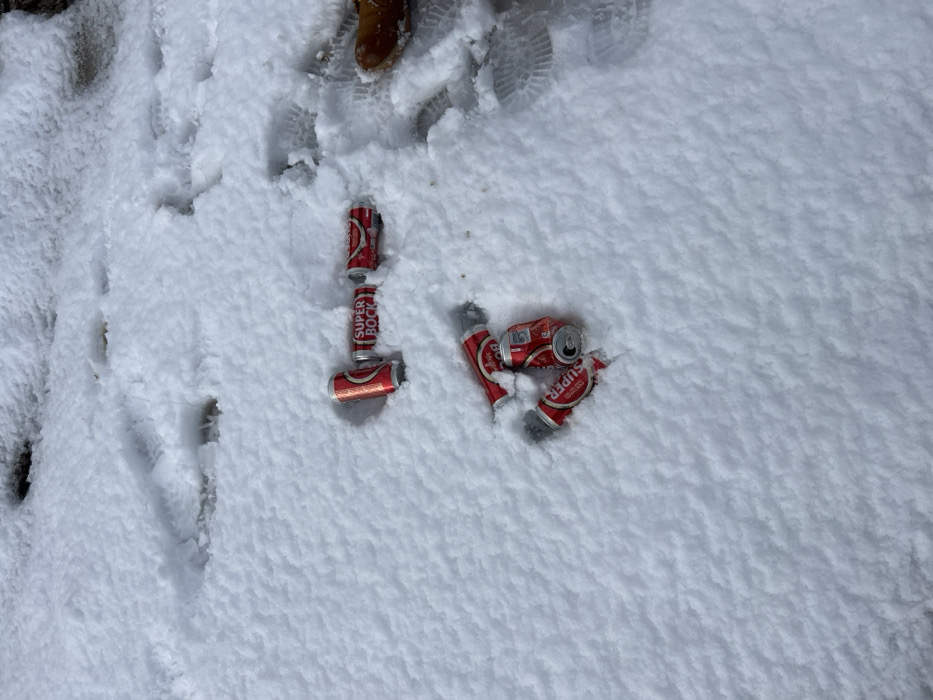 The scene depicts a snow-covered outdoor area in Vila Real, Portugal. Six red and silver "SUPER BOCK" aluminum beverage cans are present in the snow. Three cans are arranged horizontally in the mid-right portion of the frame, partially embedded in the snow. The can furthest left in this group clearly displays the "SUPER BOCK" logo. Three additional cans are grouped together in the lower-central area, appearing partially crushed and more deeply embedded. Numerous footprints are visible in the snow, indicating recent pedestrian traffic. A brown boot is partially visible on the far right edge of the frame, near some footprints. The ground is uniformly covered in fresh, white snow, with no other discernible features.
