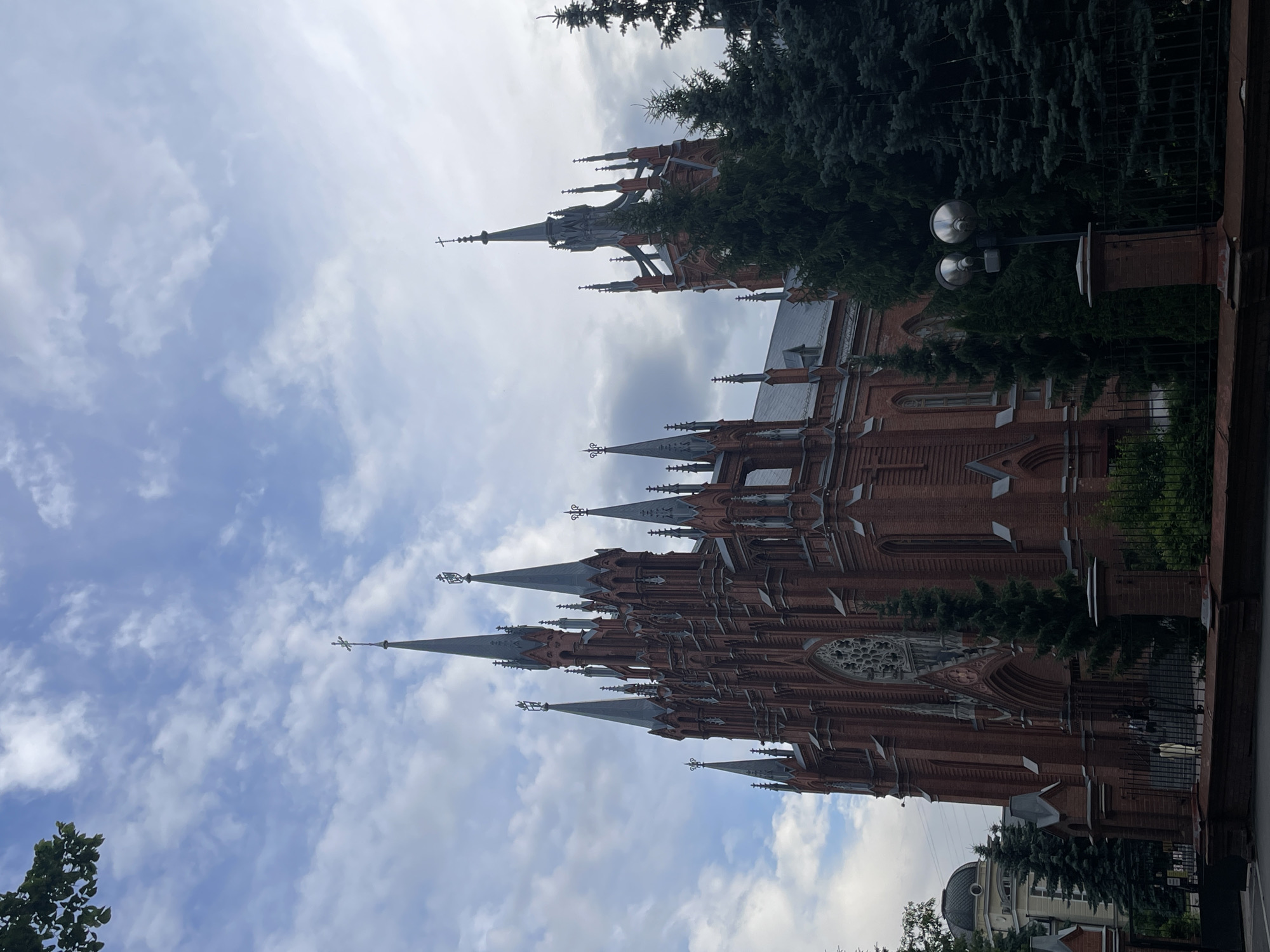 The image depicts the exterior of the Cathedral of the Immaculate Conception, a Roman Catholic cathedral located on Malaya Gruzinskaya Street in Moscow, Russia. The building is constructed of red brick in a neo-Gothic style, characterized by its tall spires, pointed arches, and ornate detailing. Several prominent spires, topped with crosses, rise towards a cloudy sky. Evergreen trees, primarily coniferous, are visible in the foreground and to the right of the cathedral, partially obscuring its lower sections. A wrought-iron fence and brick wall delineate the property line. To the left of the cathedral, a lighter-colored building with a dome is partially visible. The lighting suggests it is daytime, likely overcast.