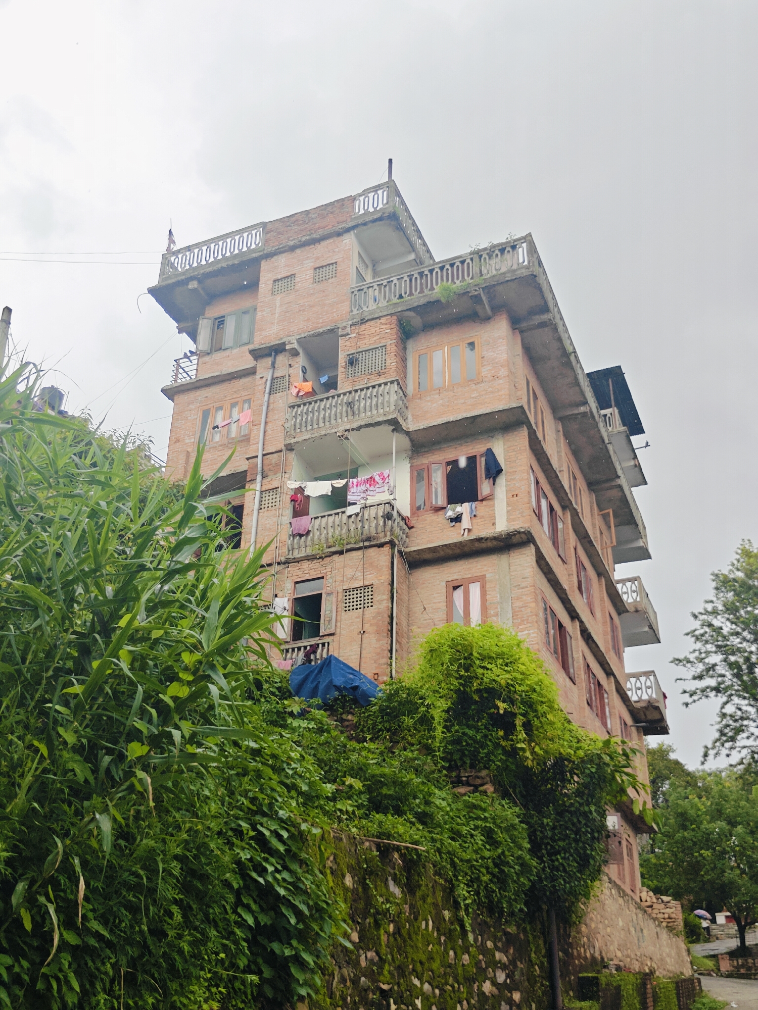 A tall, multi-story brick building is situated on a hillside in Kirtipur, Nepal. The building has several balconies with ornate railings, and laundry is hung out to dry on clotheslines. Lush green vegetation covers the hillside in front of and around the building. The sky is overcast, suggesting a cloudy or rainy day. The building's architecture appears to be typical of residential structures in the region, with exposed brickwork and concrete elements. The overall scene conveys a sense of everyday life in a hilly urban environment.