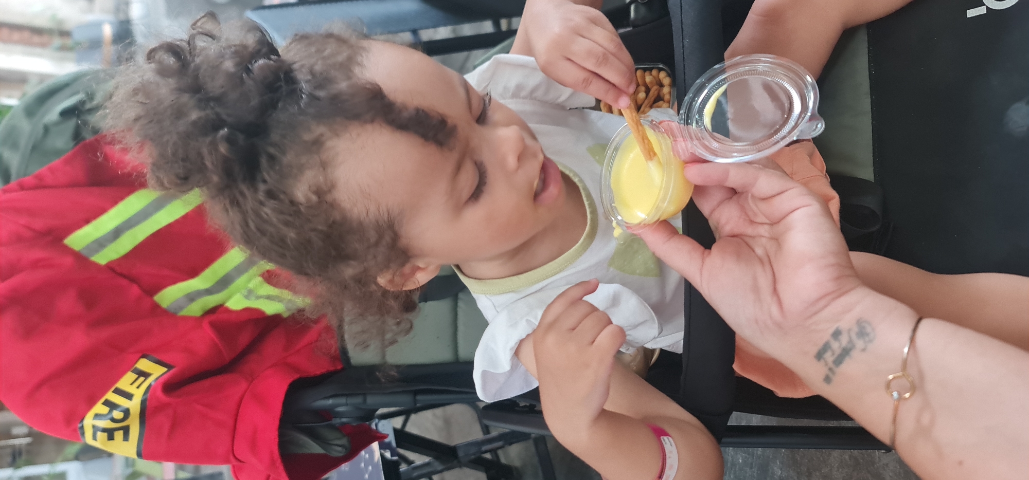 The image shows a young child with curly brown hair being fed a snack. The child is wearing a white shirt and sitting in what appears to be a stroller or wheelchair. An adult hand is holding a small clear plastic container with a yellow substance, possibly a sauce or dip, and a snack stick is being dipped into it. A red jacket with reflective yellow stripes and the word "FIRE" is draped nearby, suggesting a firefighter's uniform. The location, Đà Nẵng, Vietnam, cannot be determined from the visual information provided.