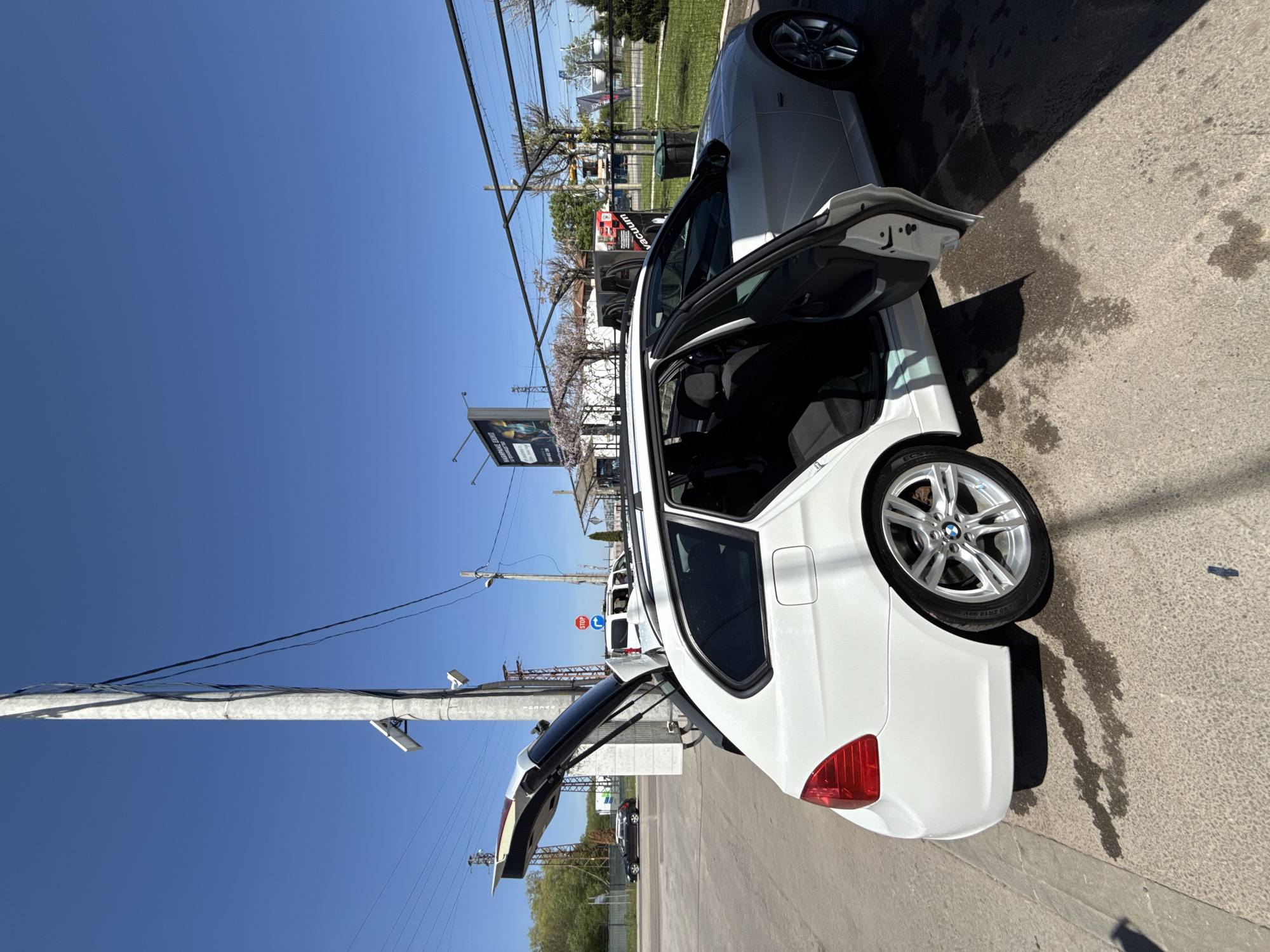 The image captures an outdoor scene, likely a car wash or detailing area, under a clear, bright blue sky in Sofia, Bulgaria. A white BMW F31 3 Series Touring (wagon) is the prominent subject, positioned on an asphalt surface. Its driver's side front door and rear tailgate are open, suggesting cleaning, detailing, or preparation. The ground shows wet patches and dark, distinct shadows, indicating strong sunlight, likely around midday.
Another white car, possibly a sedan, is partially visible in the foreground with its driver's door also open. The BMW's alloy wheels are visible, with tire sidewall text including "225/40 R18 92Y," "MICHELIN," and "ECOSPORT."
In the background, an urban or suburban landscape unfolds with multiple utility poles carrying numerous power lines. Trees, some appearing to be in blossom, line the street. A large billboard on the left displays partially legible text, possibly "ABU DHABI" and "A NEW ERA BEGINS." Further back, a red structure with the word "VACUUM" is clearly visible, supporting the car service location. A red and white "STOP" sign is also seen in the mid-ground. The scene conveys a sense of activity on a sunny day.