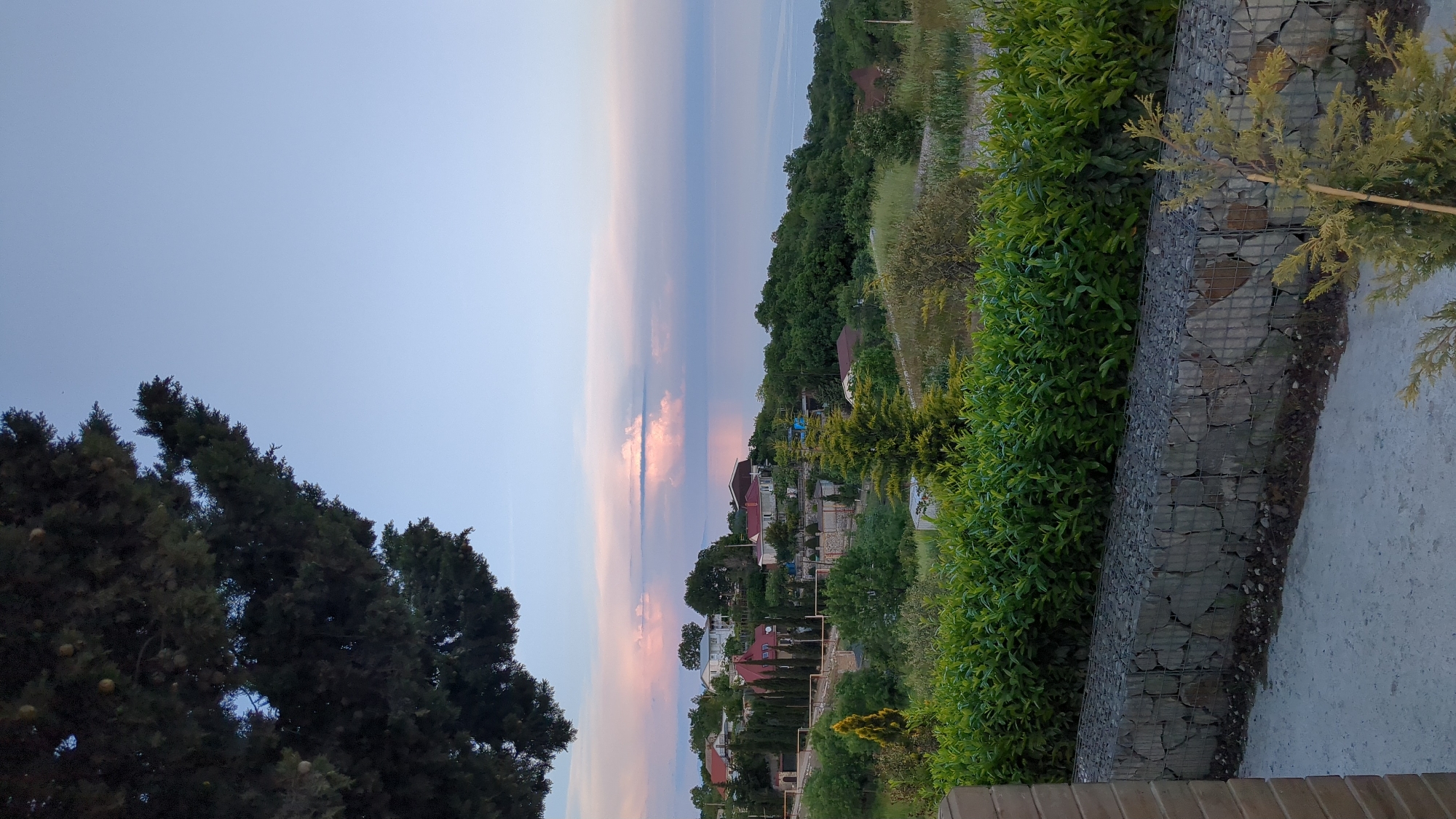 The image presents a view of Kiparisnoe, Ukraine at sunset. The sky exhibits a gradient of colors from blue to pink and peach hues where the sun's light is catching the clouds. In the middle ground, various houses with red roofs are visible amidst lush greenery. The foreground shows trees and foliage along with a stone wall and paved walkway.