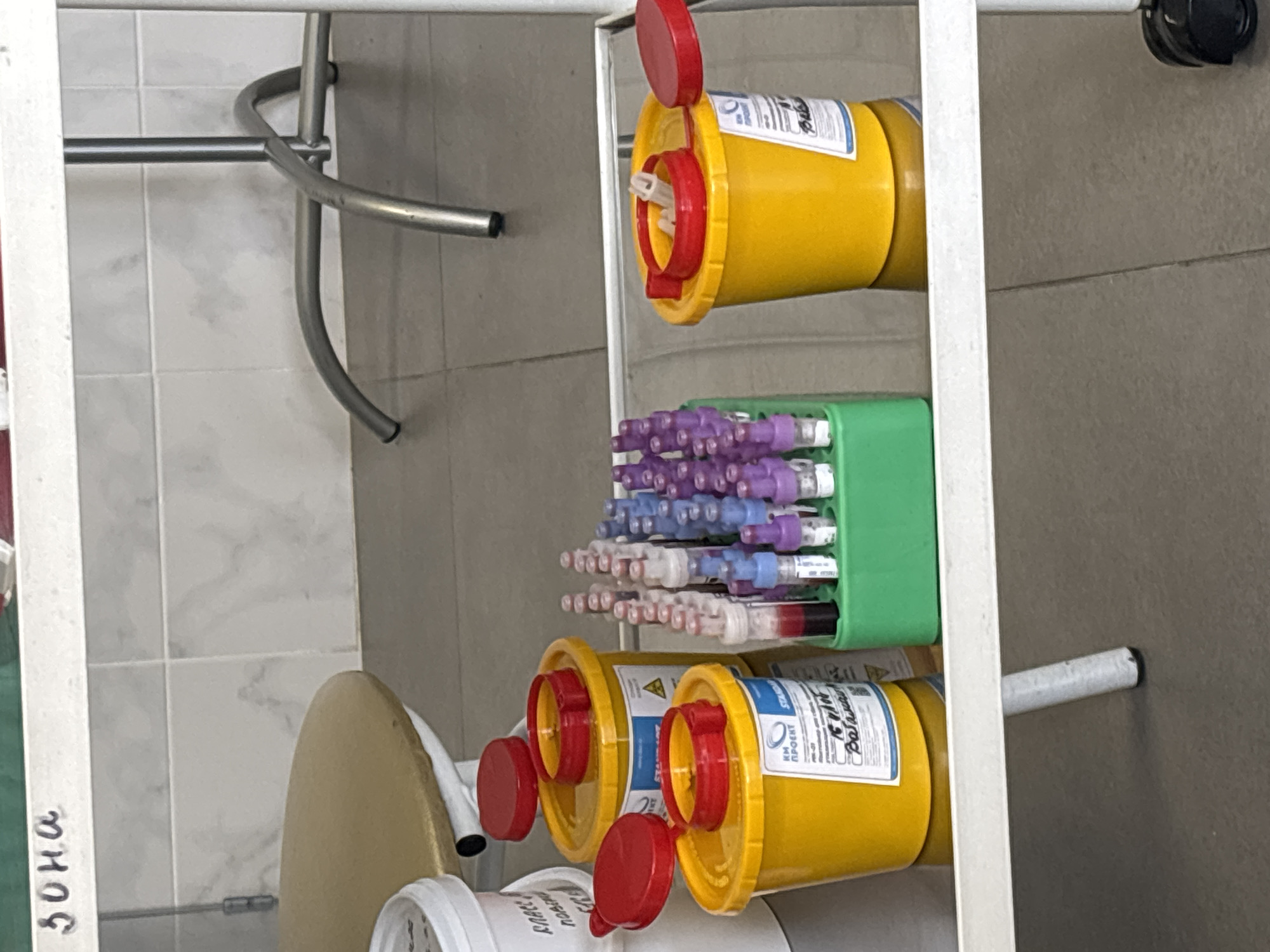 The image shows a metal rack in what appears to be a medical or laboratory setting. The rack holds two yellow sharps disposal containers with red lids and a green plastic container filled with test tubes capped with varying colors (purple, light blue, red). The containers are labeled with Cyrillic text, suggesting the location is in a Russian-speaking country. Based on the "30 HA" notation on the side of the rack, it's speculated the city is Saint Petersburg, Russia. The environment appears sterile and clinical, with tiled walls and metallic fixtures visible in the background. There is an oval yellow surface underneath the bottom container. There are no people visible in the scene. The overall impression is a functional storage setup for medical supplies and waste disposal in a medical facility.