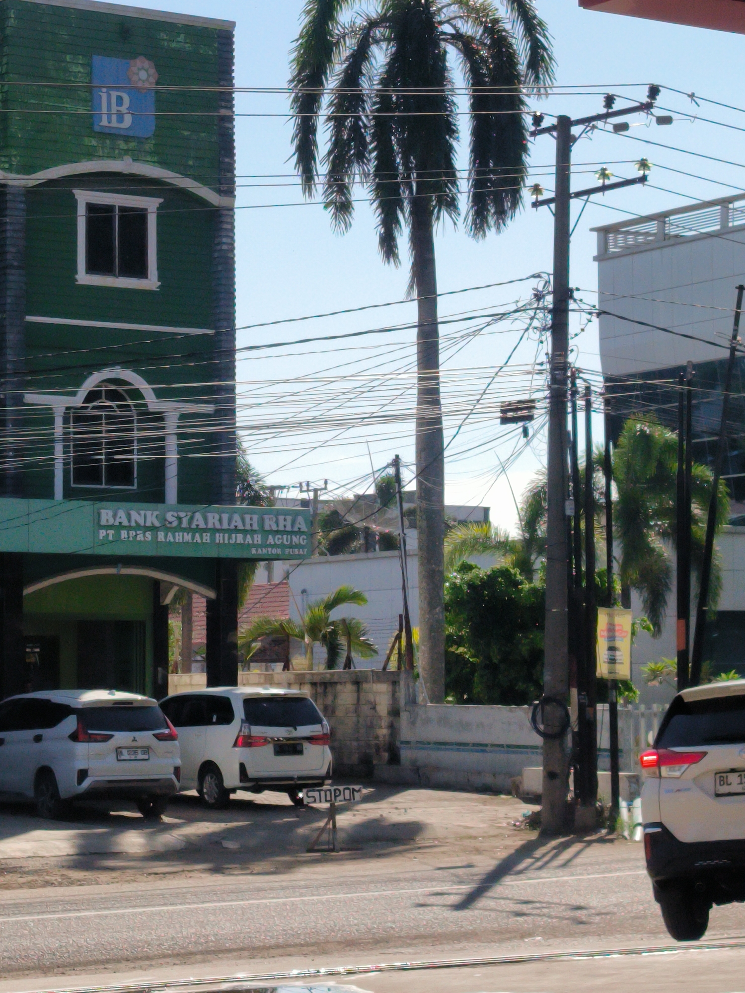 This daylight image captures a street scene in Lhokseumawe, Indonesia, characterized by urban infrastructure and tropical vegetation under a clear blue sky.
On the left, a prominent two-story building painted dark green features white architectural trim around its windows and arches. A blue square sign on the upper facade displays a logo with the letters "IB" intertwined. On the lower part of the building, a larger sign clearly reads "BANK SYARIAH RHA" with "PT BPAS RAHMAH HIJRAH AGUNG" and "KANTOR PUSAT" below it, indicating a bank branch.
Two white cars are parked in front of this green building. A third white SUV is partially visible on the far right, with its brake lights illuminated. A small, roughly made wooden sign positioned between the two parked cars reads "STOPOM".
The right half of the image is dominated by several utility poles and a dense, intricate network of overhead electrical or telecommunication wires crossing the sky. Tall palm trees are scattered throughout the scene, one prominent on the far left, and several others behind the utility poles, along with other green foliage. A modern white building with multiple levels is visible in the background on the right.
The paved road in the foreground shows reflections, suggesting a possible wet surface. Bright sunlight casts distinct shadows from the utility poles and vehicles, indicating a sunny day. License plates on the vehicles, such as "BL 1398 QQ" and "BL 18...", are visible, which are characteristic of Aceh province in Indonesia, aligning with the Lhokseumawe context. No people are discernible in the frame, creating a relatively quiet street depiction.