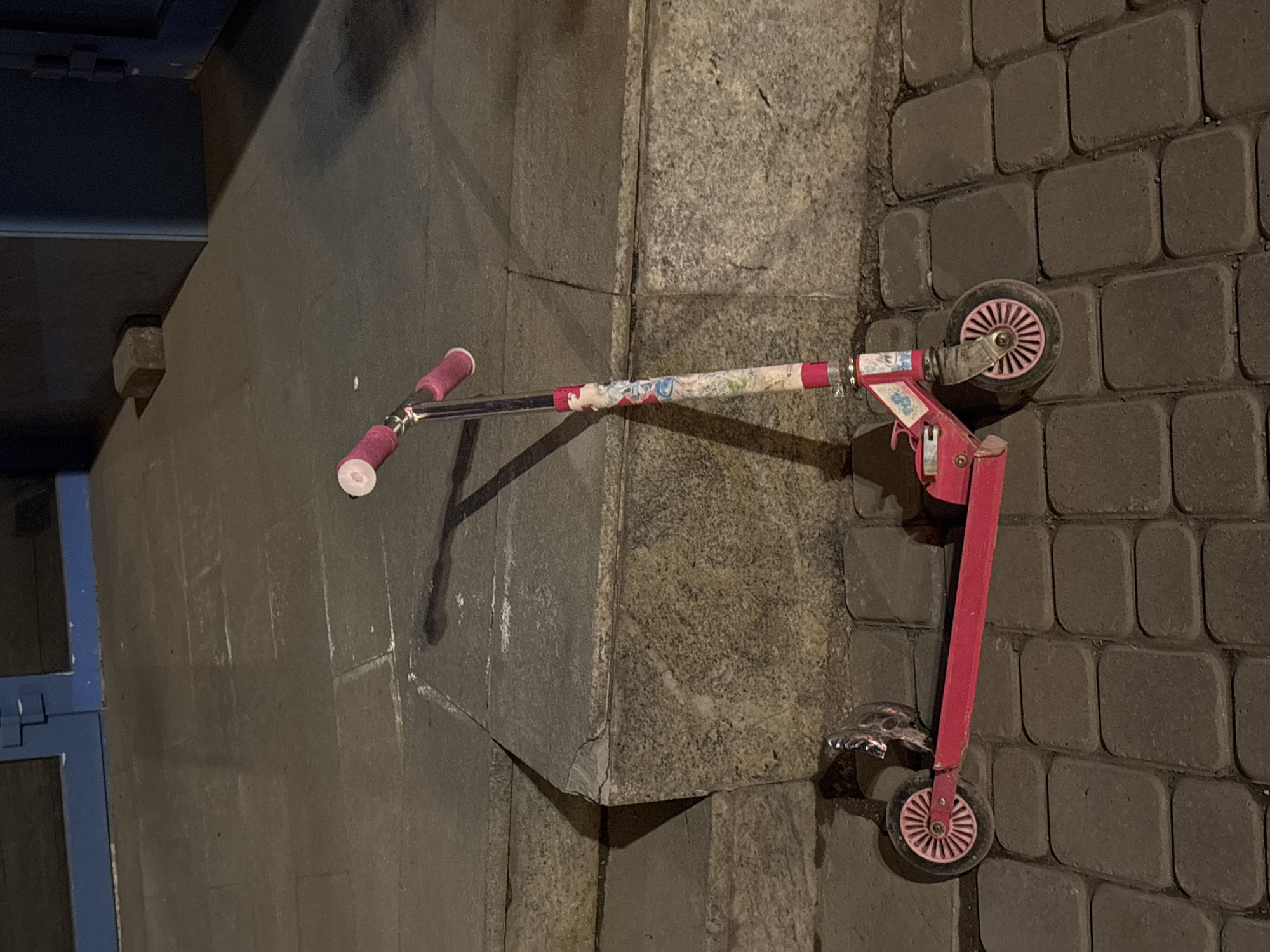 A pink scooter rests on a sidewalk constructed of dark grey paving stones. The scooter is leaning against a concrete structure. The setting appears to be an urban area, as indicated by the paving and presence of a doorway/entryway behind the scooter. While this information cannot be definitively ascertained from the image alone, the information included in the prompt states this scene is in Krasnoyarsk, Russia.