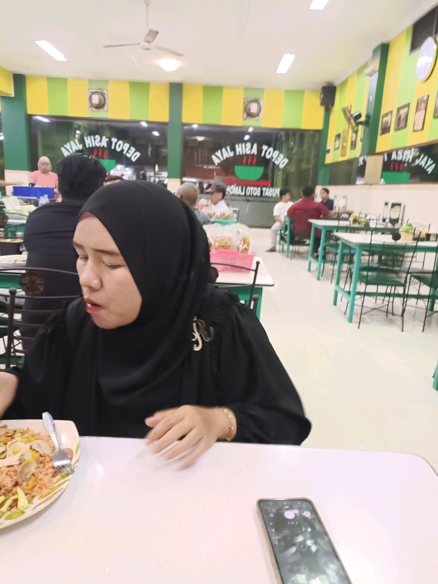A woman in a black hijab and black top is seated at a table, looking down and eating from a plate of food. A cell phone is on the table in front of her. The restaurant appears to be busy with other patrons and tables set up. Signs on the wall read "AYAL HISA TOPQ" and "ASIH JAYA". The walls are painted with yellow and green stripes. The overall environment suggests a casual dining establishment. The location is Dapur, Indonesia.