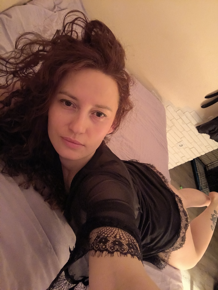 A female subject is depicted indoors, lying on a bed with light lavender bedding. Her head is positioned to the upper left of the frame, with her body angled away towards the lower right. She has long, curly, reddish-brown hair. Her left eye is slightly larger than her right, and her lips are slightly parted. She is wearing a sheer black robe or nightgown with black lace trim on the sleeves and hem. A dark, possibly floral or leafy, tattoo is visible on her lower left leg, near the ankle. The subject looks directly towards the camera with a neutral expression. In the background to the right, a beige wall is visible above a section of white rectangular tiling. Further right and lower, a dark, multi-barred object, possibly a pet crate, rests on a dark floor surface. The lighting is warm and ambient. There are no visual cues within the image to specify a location in the city of Saint Petersburg, Russia.