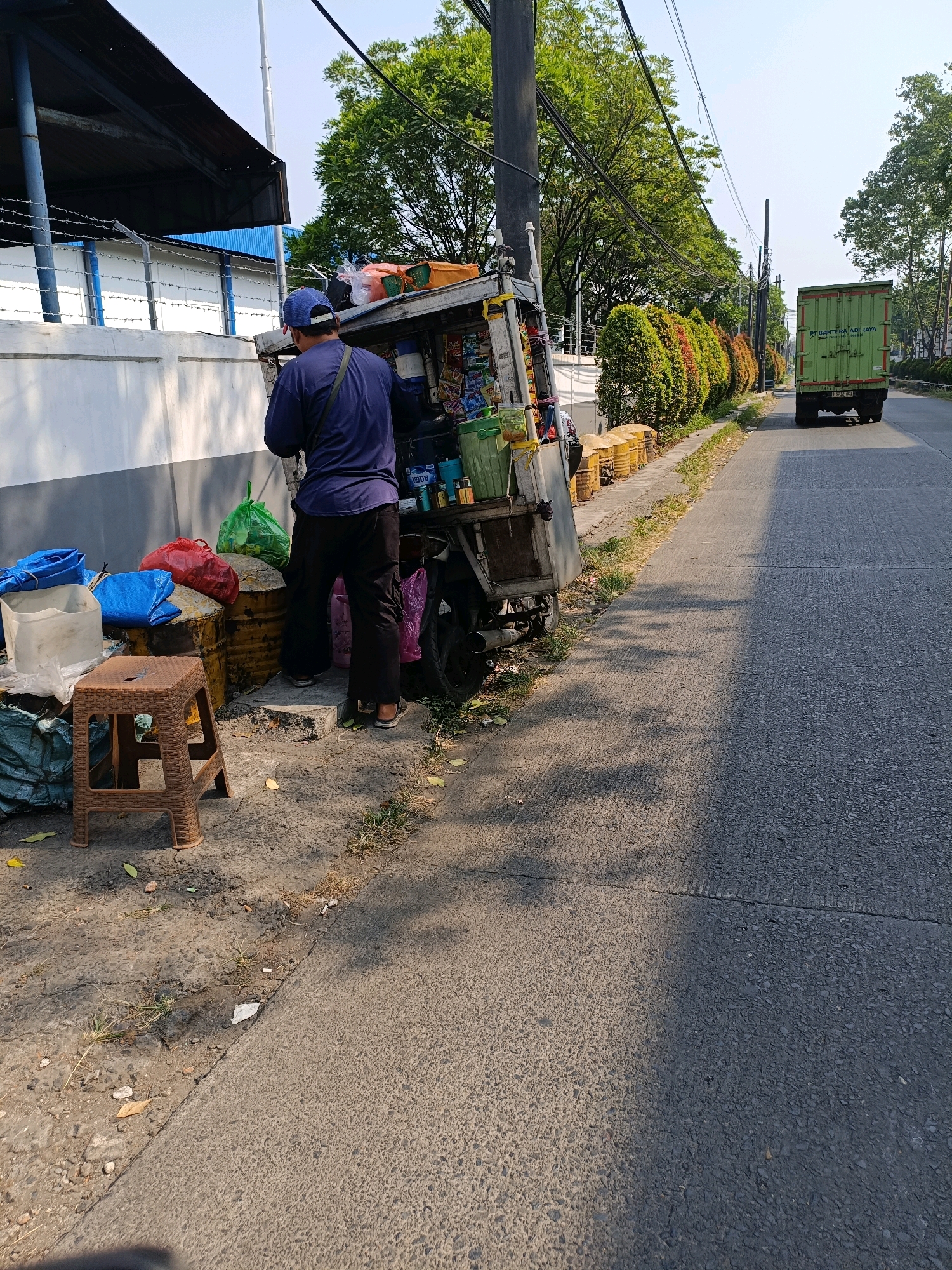 The image depicts a street scene in Tangerang, Indonesia. In the foreground, a person wearing a dark blue shirt and a blue baseball cap stands beside a mobile cart, which appears to be a food or goods vendor. The cart is laden with various items, including bottles, packaged goods, and containers. Next to the cart, several stacked items, including what appear to be oil drums and bags of various colors, are visible. A small, brown plastic stool is placed on the ground. The cart is parked on a paved surface next to a sidewalk bordered by a low wall and a building. The wall has barbed wire along the top. Further down the street, a green box truck is driving away from the camera. The environment is characterized by a paved road, sidewalk, vegetation including trees and manicured bushes, and electrical wires overhead. The sky is clear and blue. The overall impression is of a typical urban street with commercial activity.