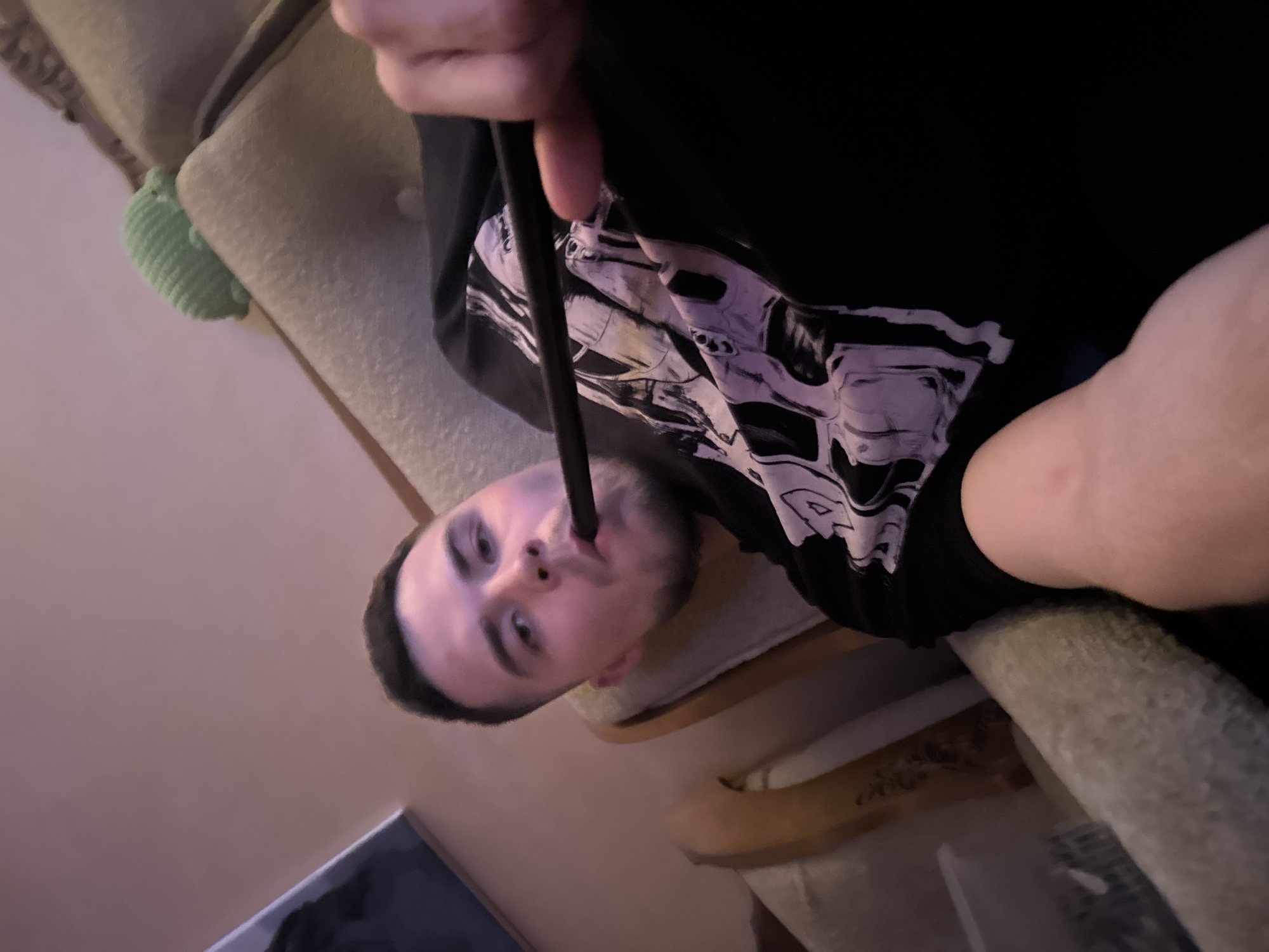 One male individual is depicted indoors, reclining in an upholstered armchair with visible wooden framework. The individual, appearing to be an adult with dark hair, a light complexion, and facial stubble, is wearing a black t-shirt with a light-colored graphic design. He is holding a black tubular object, consistent with a hookah hose, to his mouth, indicating the act of smoking or preparing to smoke. His right hand is visible holding the object.
The background consists of a plain, light-colored wall. A small, ribbed, light green, elongated object, possibly a toy or decorative item, rests on the back of the armchair to the right of the individual. A dark-framed object, possibly a picture or mirror, is partially visible on the wall to the left. The ambient lighting is dim and warm, consistent with artificial indoor illumination. The perspective suggests a low-angle shot, possibly a self-portrait. There are no discernible elements within the image to confirm the specific location as Perm, Russia.