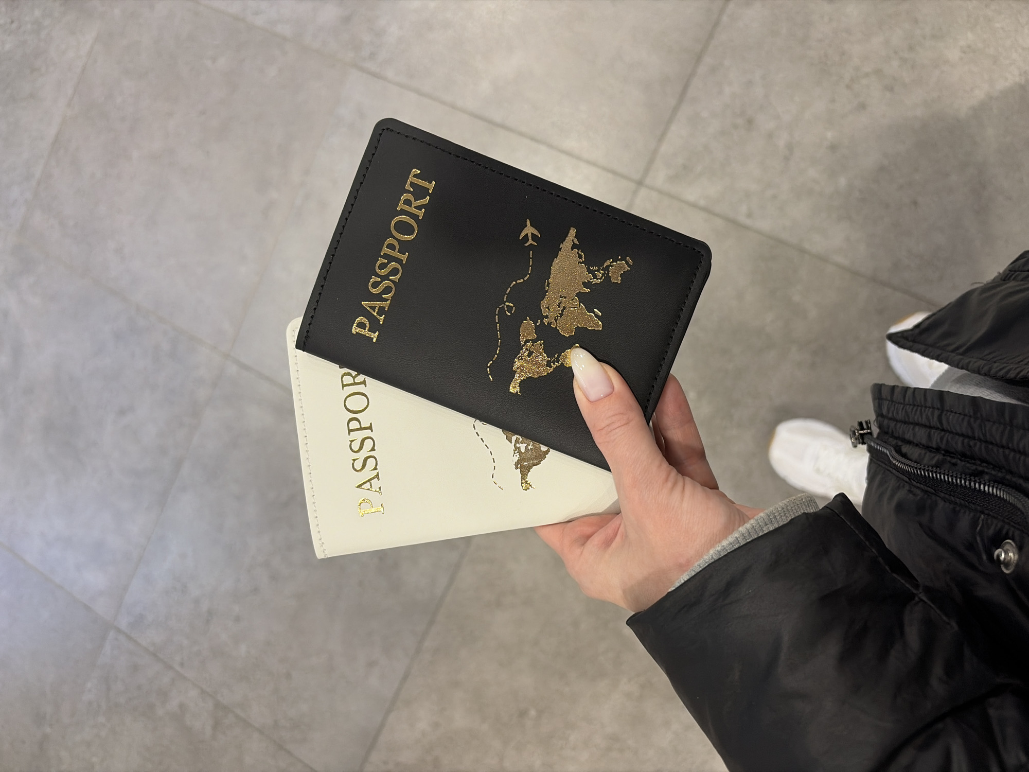A person's right hand holds two passport covers, one black and one cream-colored, above a light grey tiled floor. The black cover is fully visible, featuring "PASSPORT" in gold lettering along with a gold world map design and a dashed airplane travel path. The cream-colored cover, partially obscured behind the black one, also displays "PASSPORT" in gold. The hand has manicured fingernails with white tips. The individual is wearing a black quilted jacket and a grey long-sleeved garment, with white athletic shoes visible on their right foot. The scene takes place in the city of Boldino, Russia.