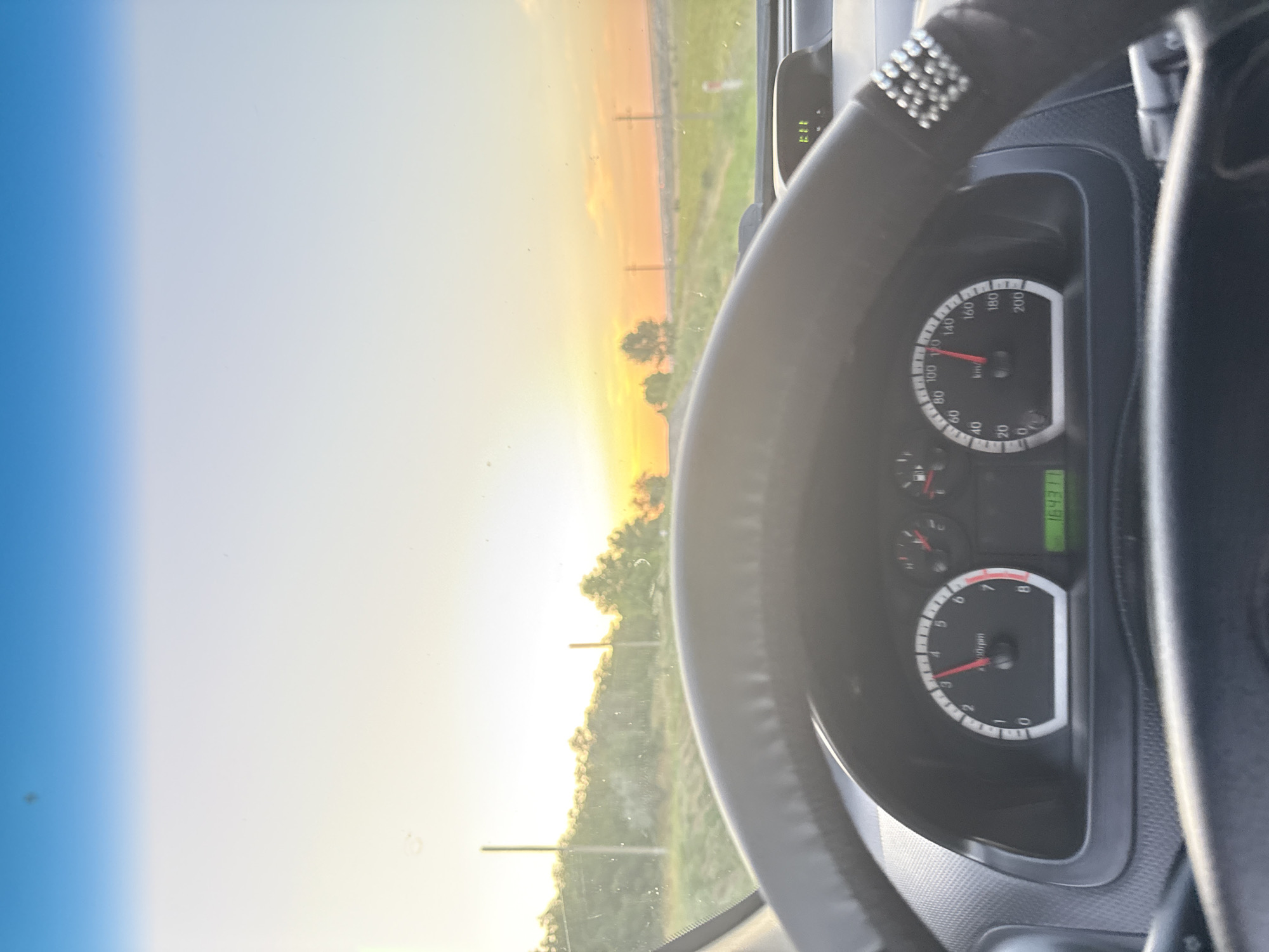 The image is taken from the driver's perspective inside a car, looking out the windshield at a sunset. The dashboard is visible in the foreground, with the steering wheel slightly off-center. The speedometer shows the needle pointing around 120 km/h, and the odometer reads 164371. The sky is a gradient of blue to bright orange and yellow, indicating sunset. Outside the car, there are trees and poles visible, with a grassy field stretching into the distance. The lighting suggests a rural or semi-rural setting. The car appears to be in motion. The specific location is identified as Cioc-Maidan, Moldova.